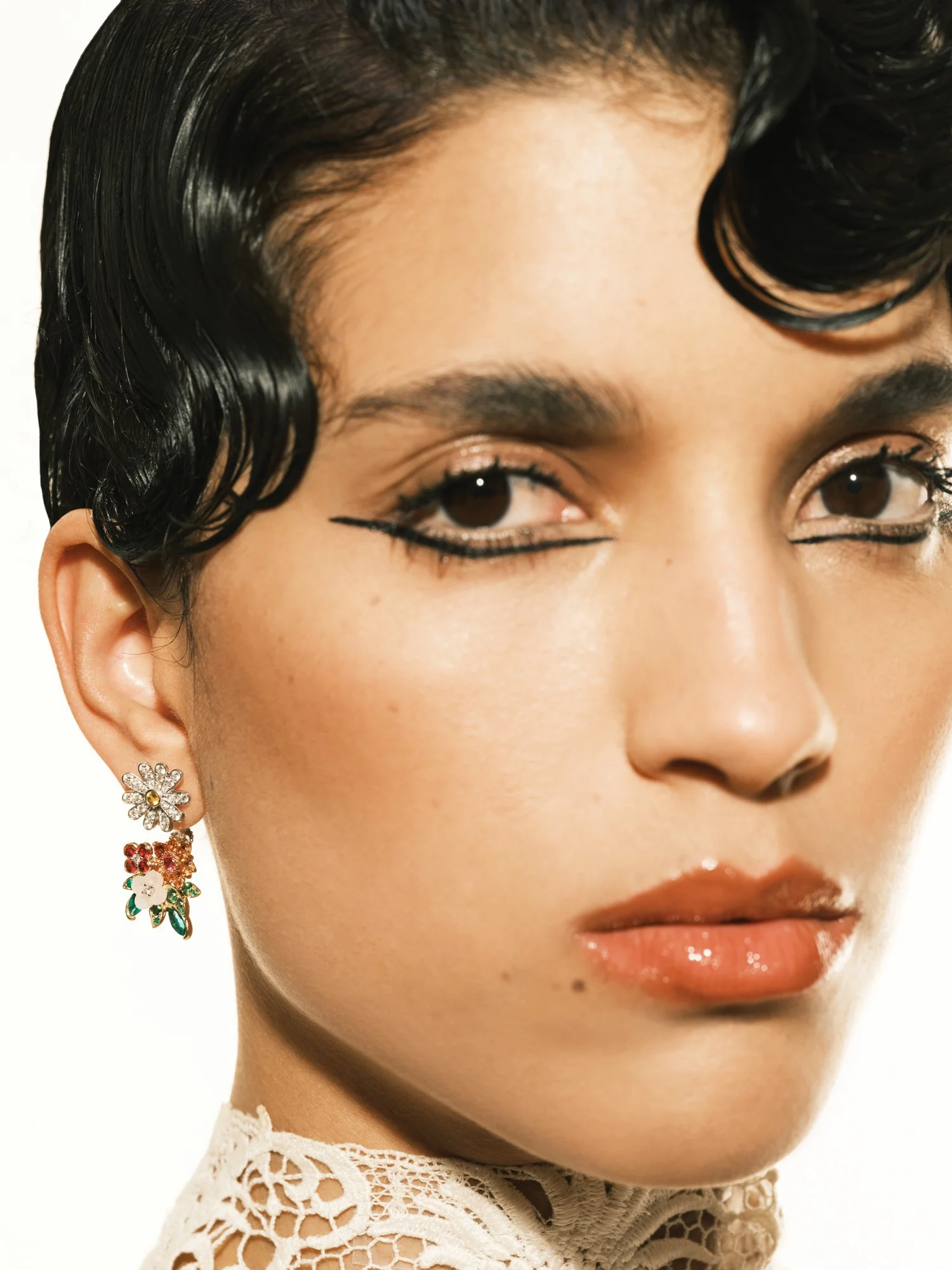 Close-up of a woman with short black wavy hair, wearing floral earrings, with makeup including black eyeliner, nude lips, and wearing a lace top.