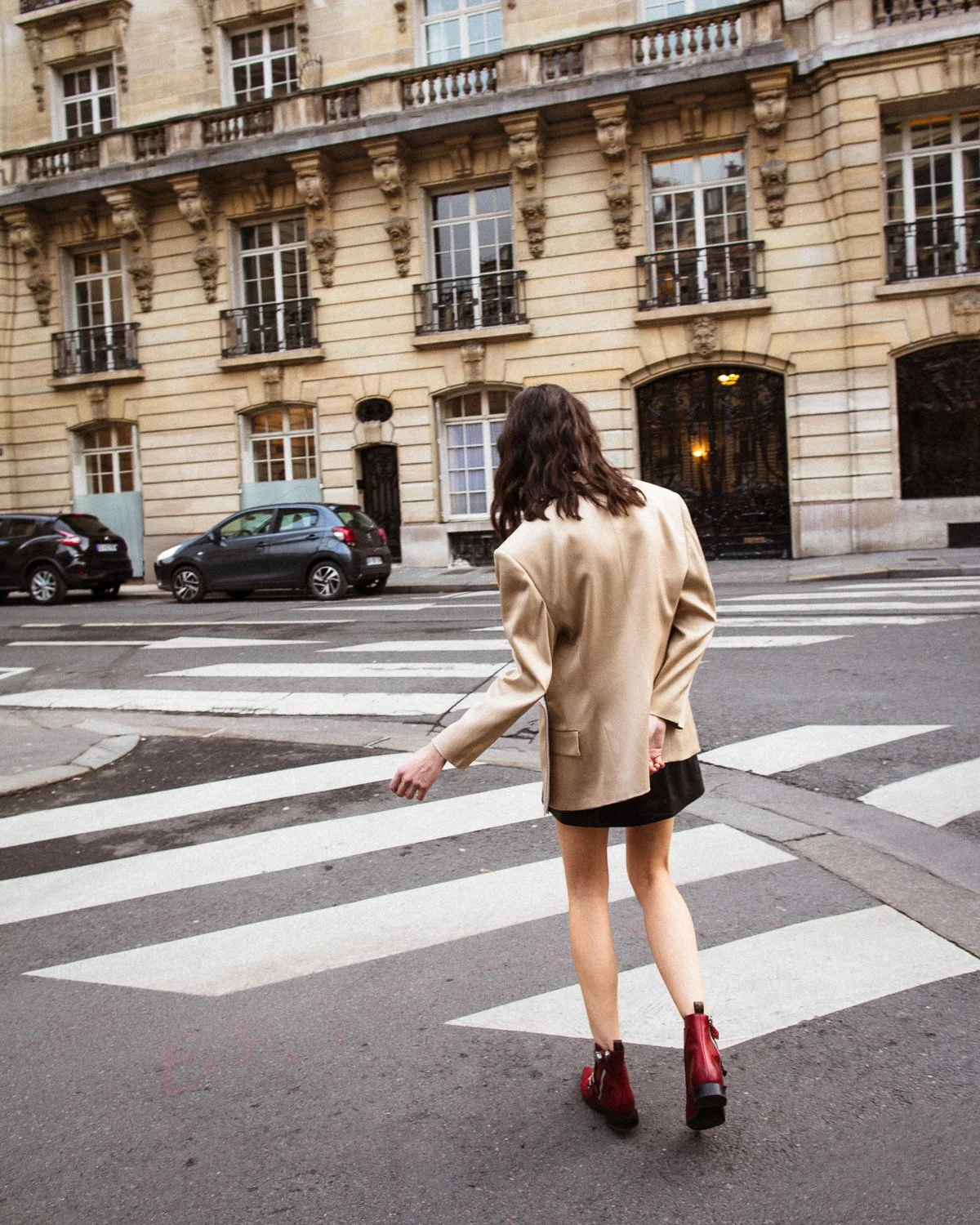 A woman crossing a city street at a crosswalk, wearing a beige blazer, black skirt, and red boots, with her back to the camera, in front of an old building with multiple windows and small balconies.