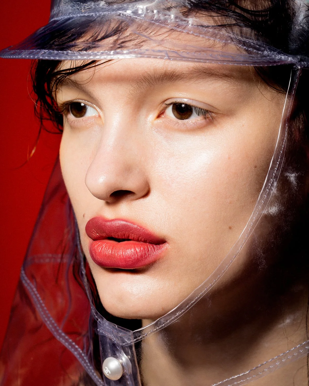 Close-up of a young woman's face wearing a transparent plastic hat with a pearl button, red lipstick, and smooth skin, with a red background.
