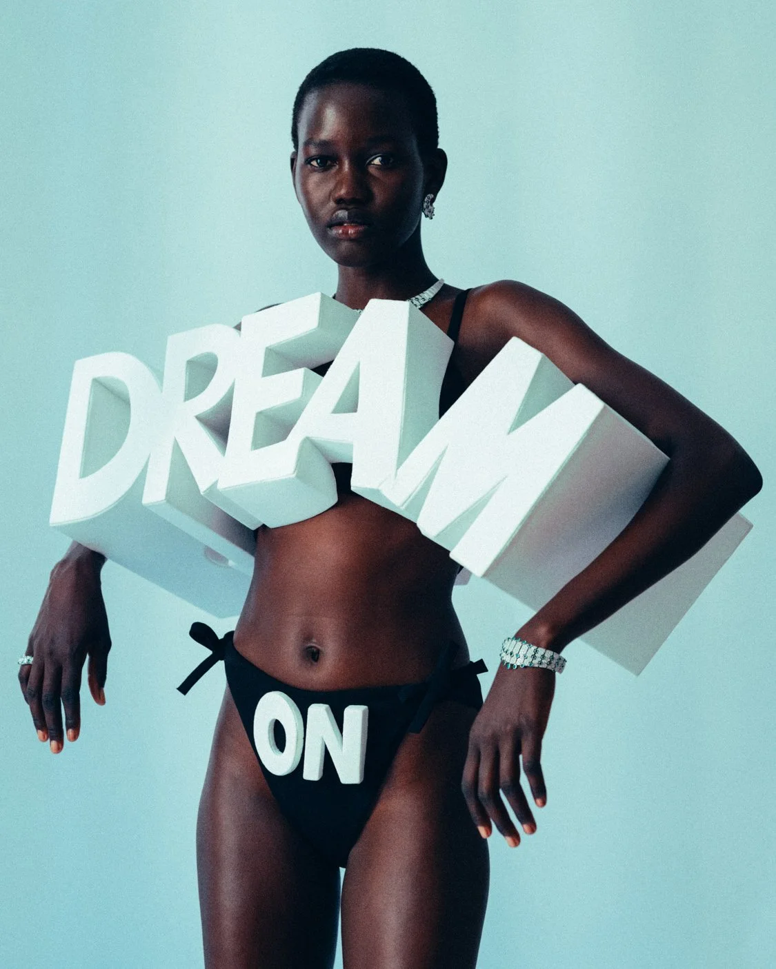 A woman wearing black underwear with white letters that say 'DREAM' on her chest and 'ON' on her underwear, holding a large 3D white 'DREAM' sign in front of her chest.