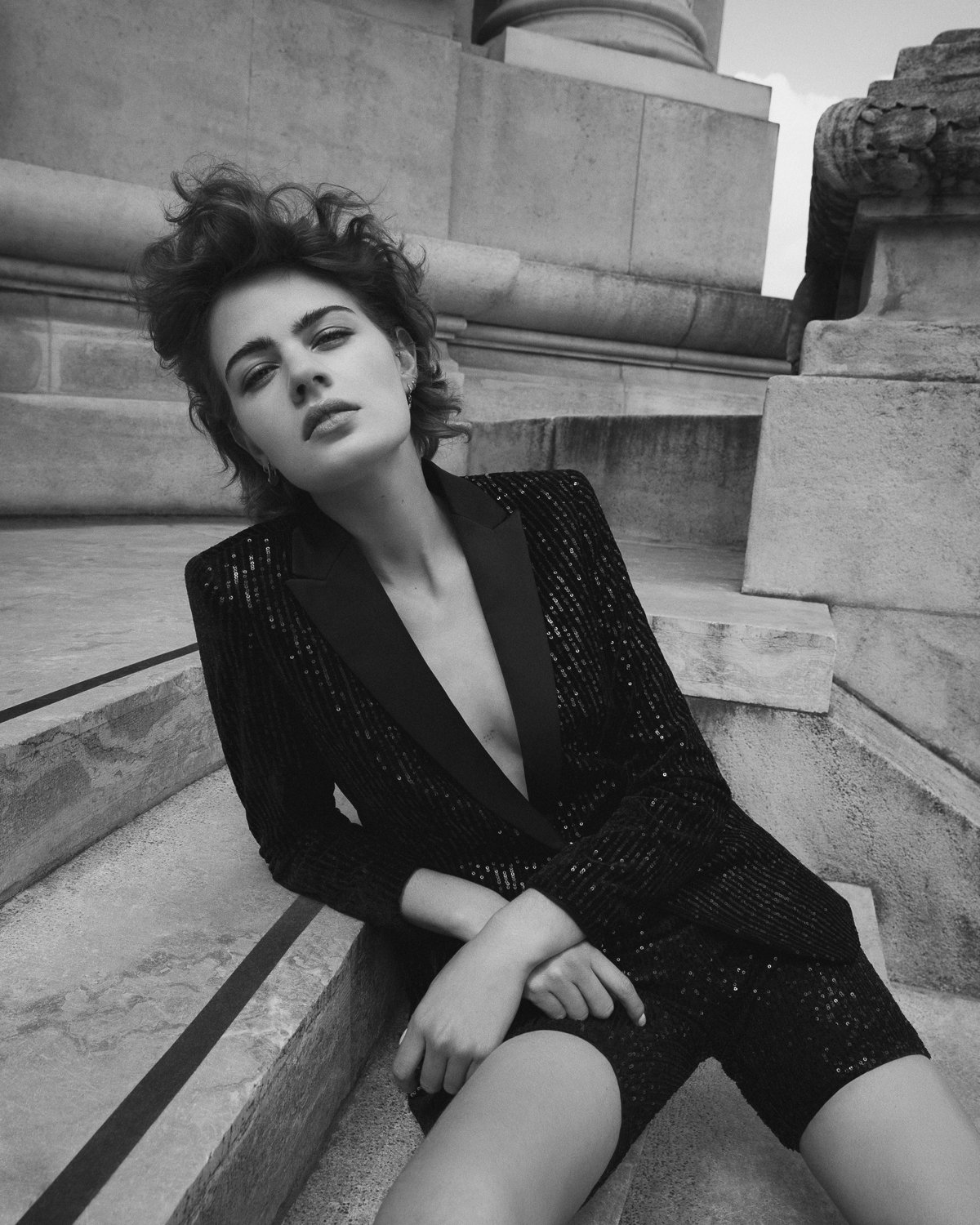 A young woman with curly hair and bold makeup, wearing a black sequin blazer and shorts, sitting on stone steps outdoors, with an intense expression, in black and white.