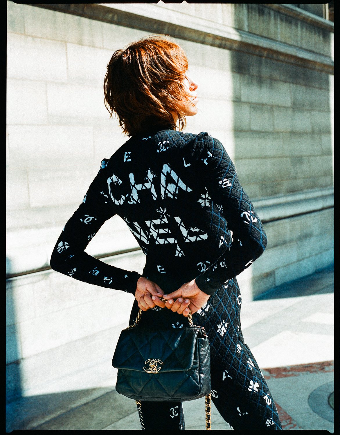 A woman with short, reddish-brown hair standing outdoors in front of a stone wall, wearing a black and white patterned outfit and holding a black quilted handbag with a chain strap behind her back.