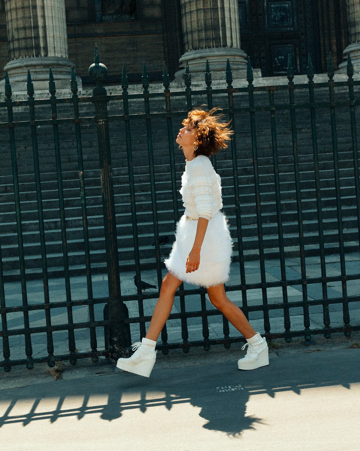 A woman walking past a black iron fence and steps in front of a historic building. She has short, curly hair and is wearing a white layered sweater, a fluffy white skirt, white socks, and white platform shoes. She appears to be enjoying sunny weather