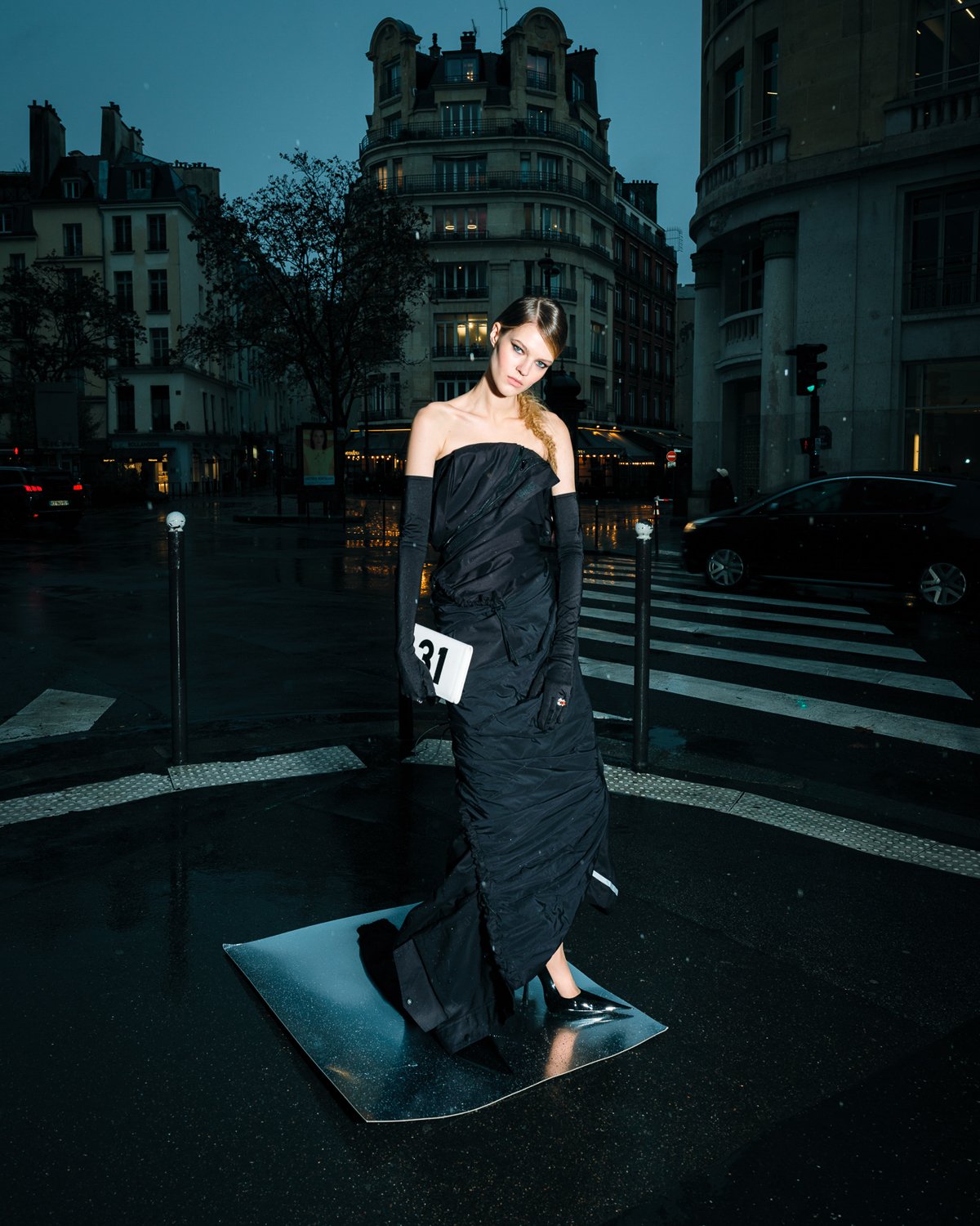 Fashion model in a black gown standing on a reflective surface in an urban street at night.