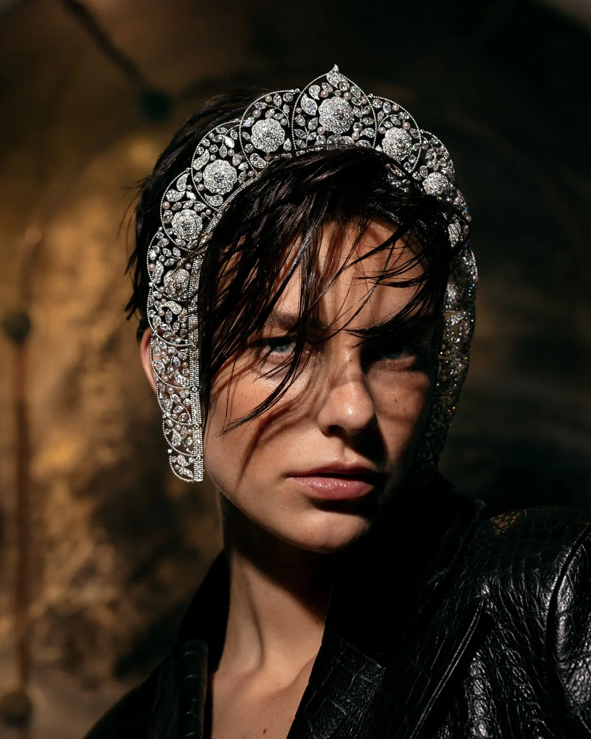 Close-up of a woman wearing a shiny, ornate tiara with floral patterns, dark wet hair, and a black leather jacket, with a dark and blurred background.