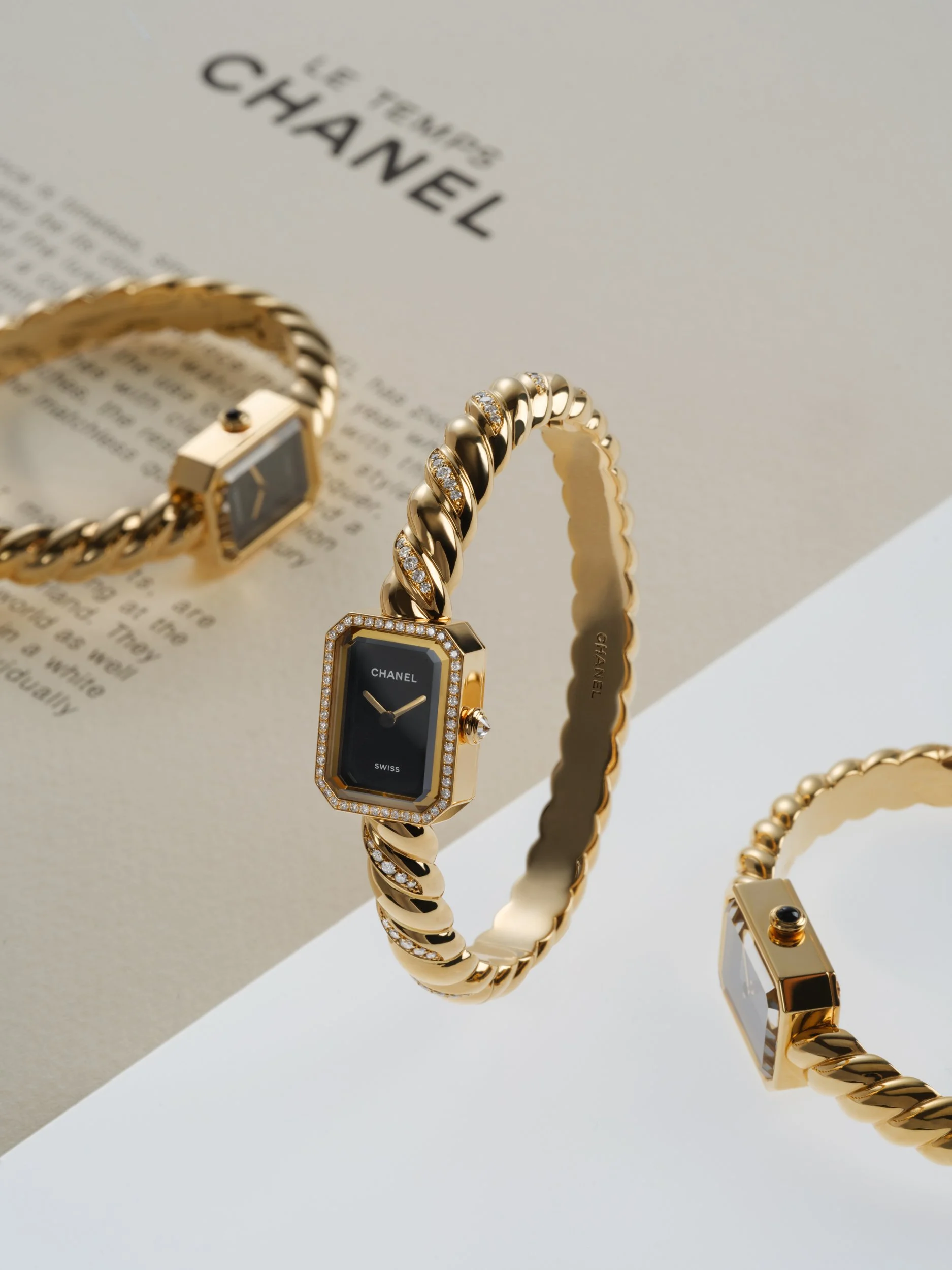 Three gold Chanel wristwatches with black faces and diamond accents displayed on a white and beige background.