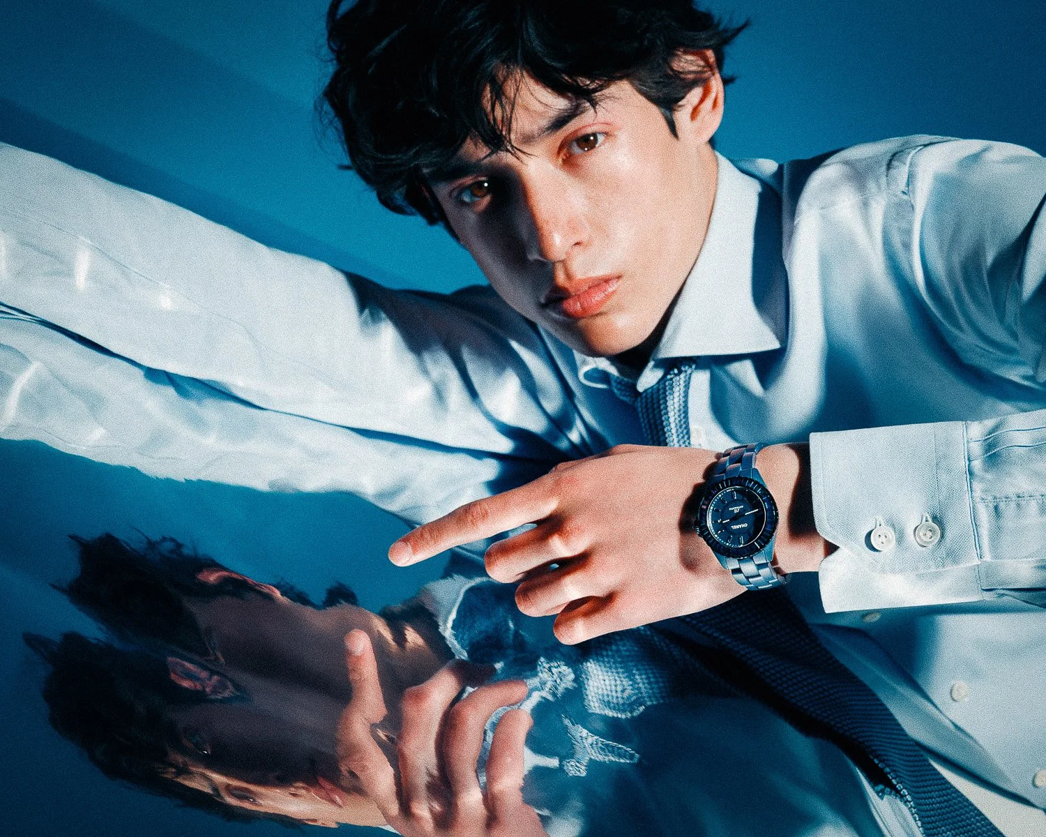 A young man with dark curly hair lying on a reflective surface, wearing a light-colored shirt, pointing at a wristwatch, with his reflection visible.
