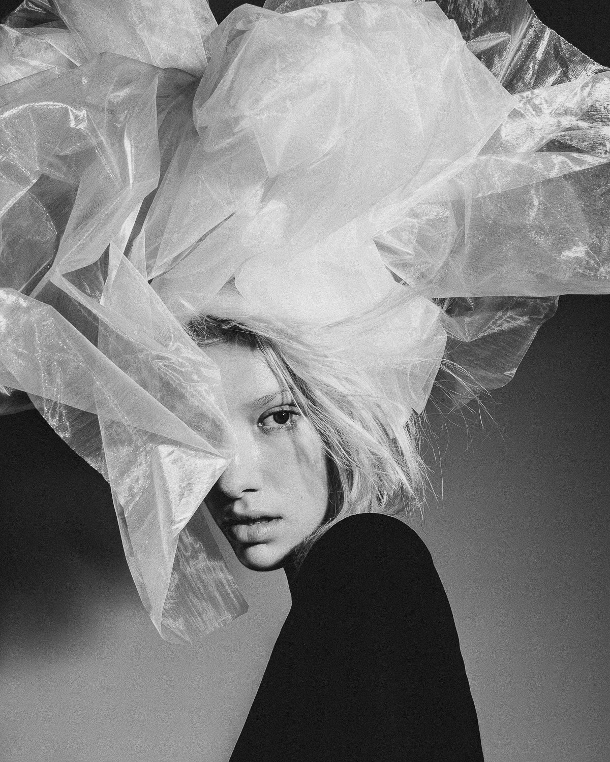 Black and white portrait of a woman with light-colored hair, partially covered by translucent plastic fabric, looking towards the camera with an intense expression.