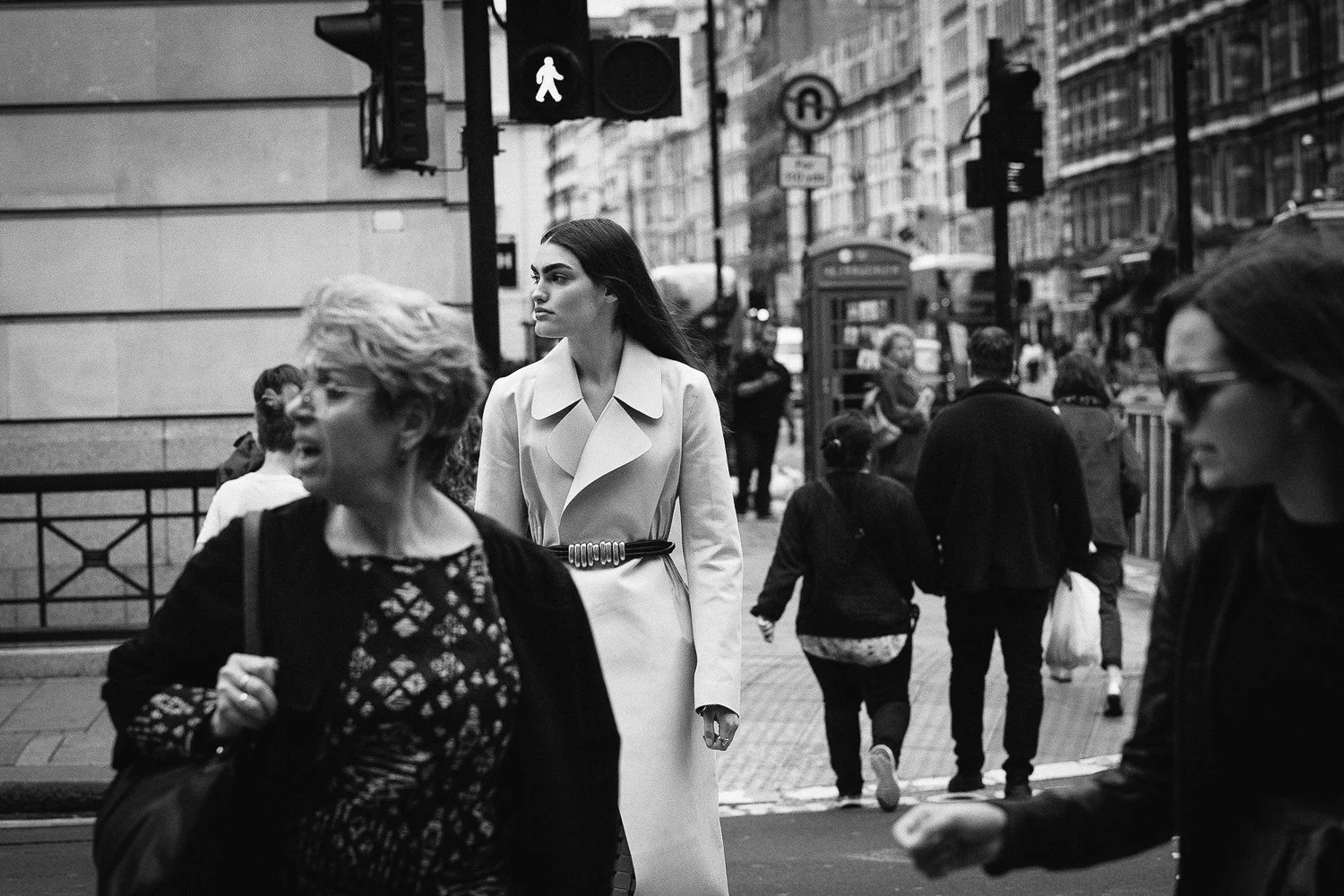 A busy city street scene in black and white showing pedestrians walking past a woman in an elegant white coat, with various other people and city buildings in the background.