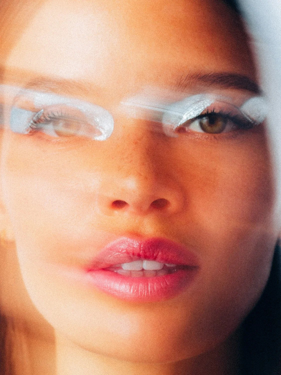 A close-up of a woman with on her face with eye makeup, including silver eyeshadow and white eyeliner. She is looking slightly to the side, with parted lips showing some teeth.