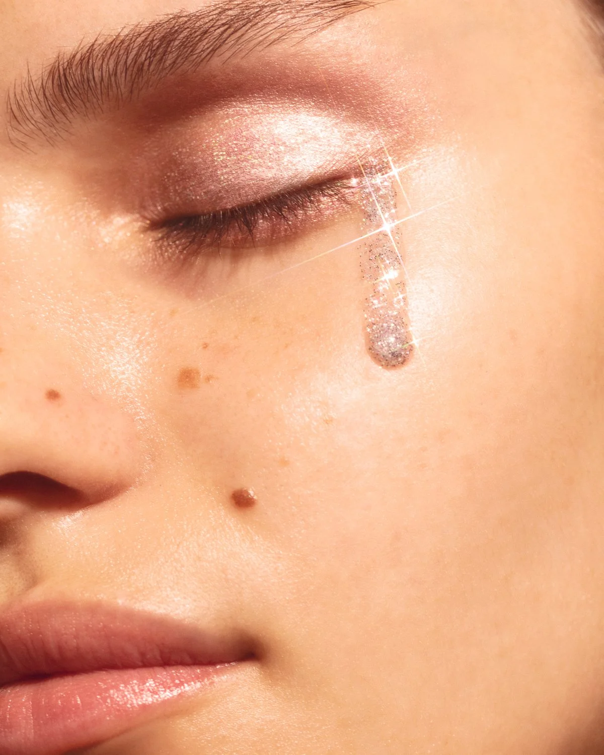 Close-up of a woman's face showing makeup with glitter eyeshadow, lips, and a tear-shaped jewel beneath her eye, with natural skin texture and moles.