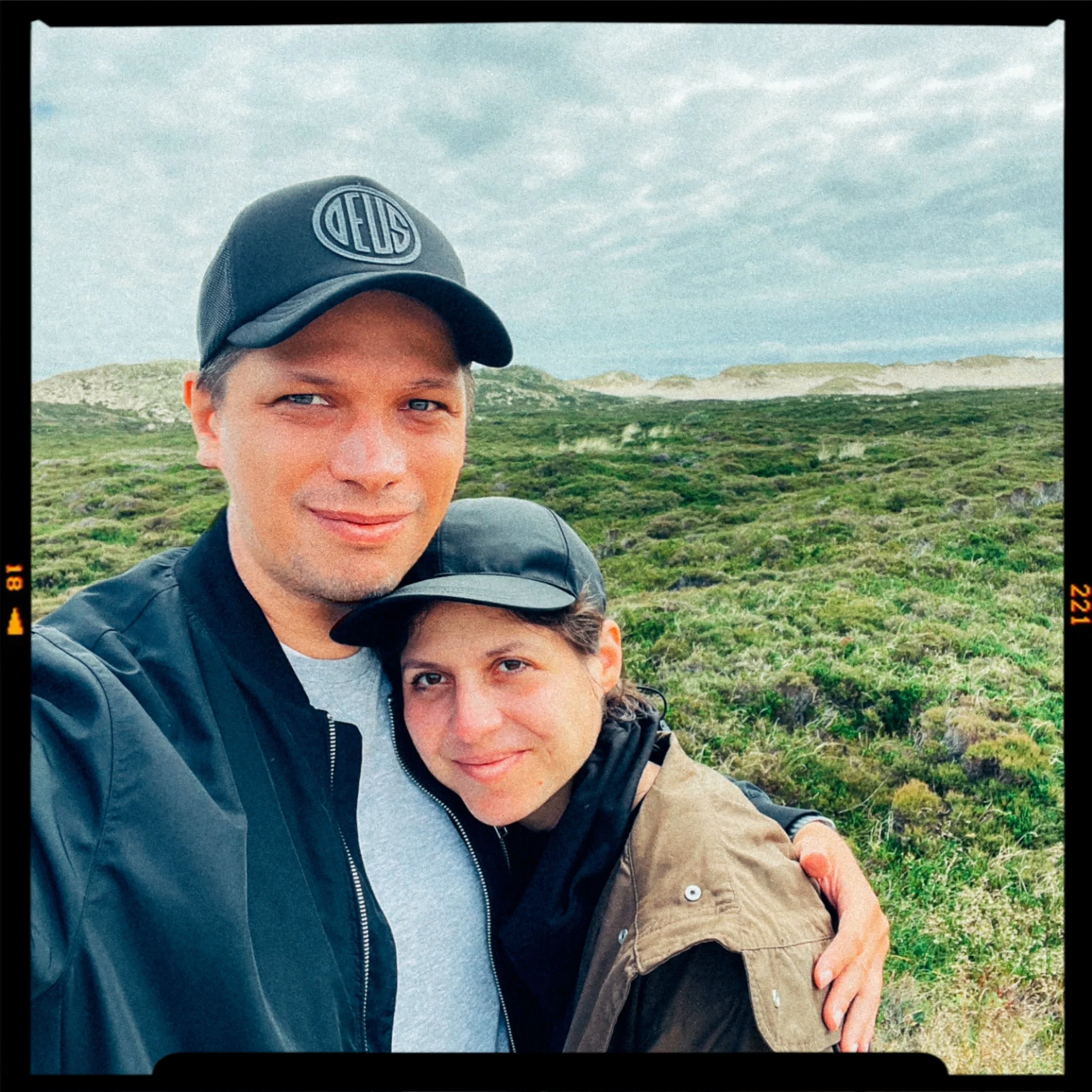 A smiling man and woman taking a selfie outdoors in a green, hilly landscape with cloudy skies.