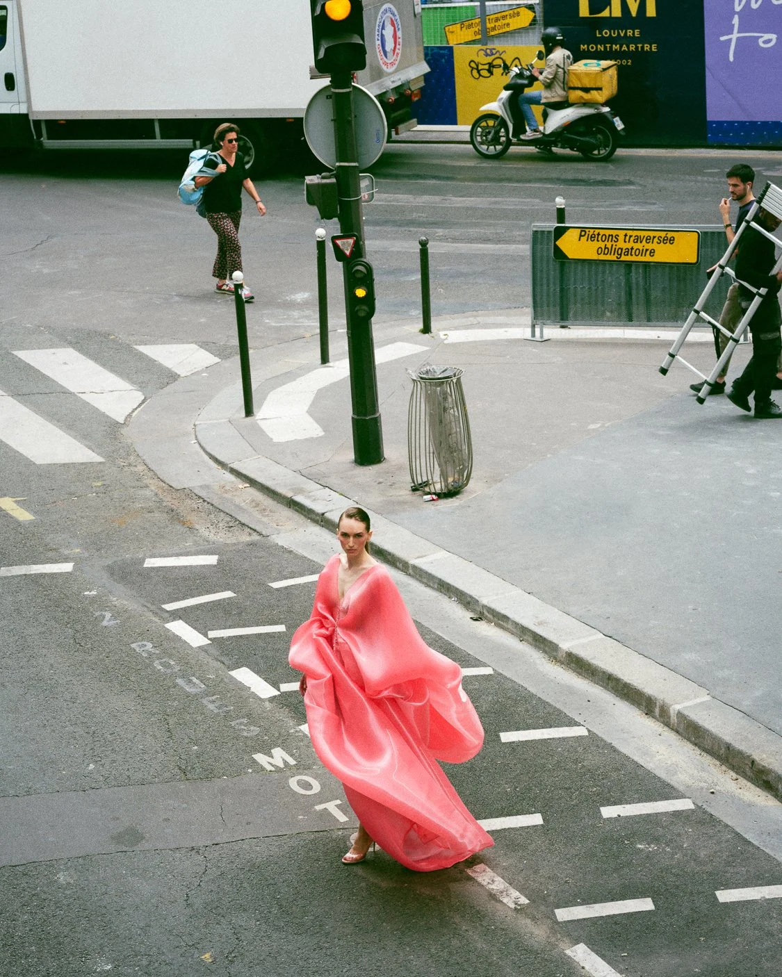 A female model wearing a flowing pink dress stands in the intersection of a Paris street, with crosswalk lines visible on the road. In the background, pedestrians, a delivery scooter, and a truck are seen, along with a yellow sign in French that read