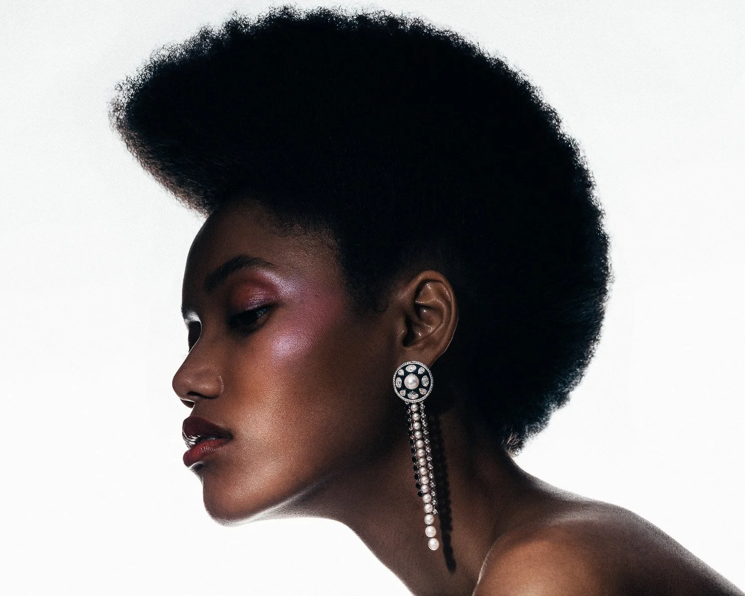 Profile of a woman with dark skin, natural hairstyle, wearing large, ornate earrings, with makeup highlighting her cheekbones and eyelids.