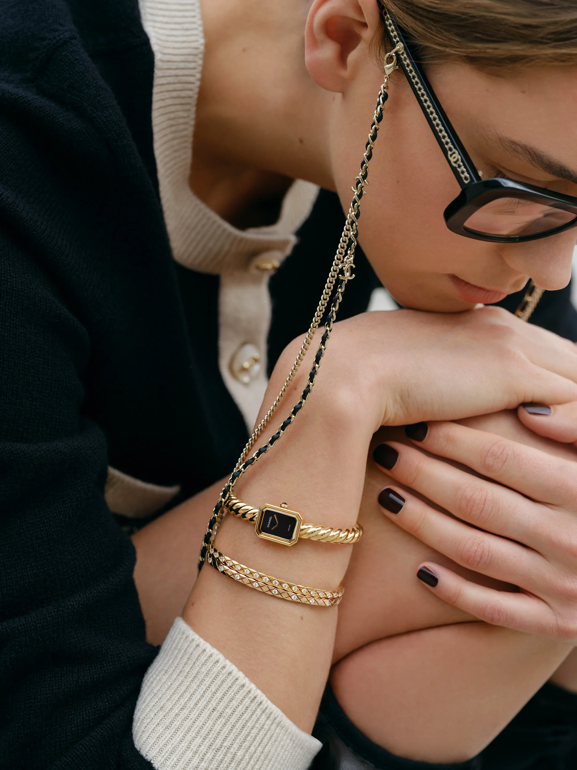 A woman wearing glasses and jewelry, including a black and gold watch, gold and black chain bracelet, and a gold with diamond bracelet on her wrist, with her hand on her shoulder.