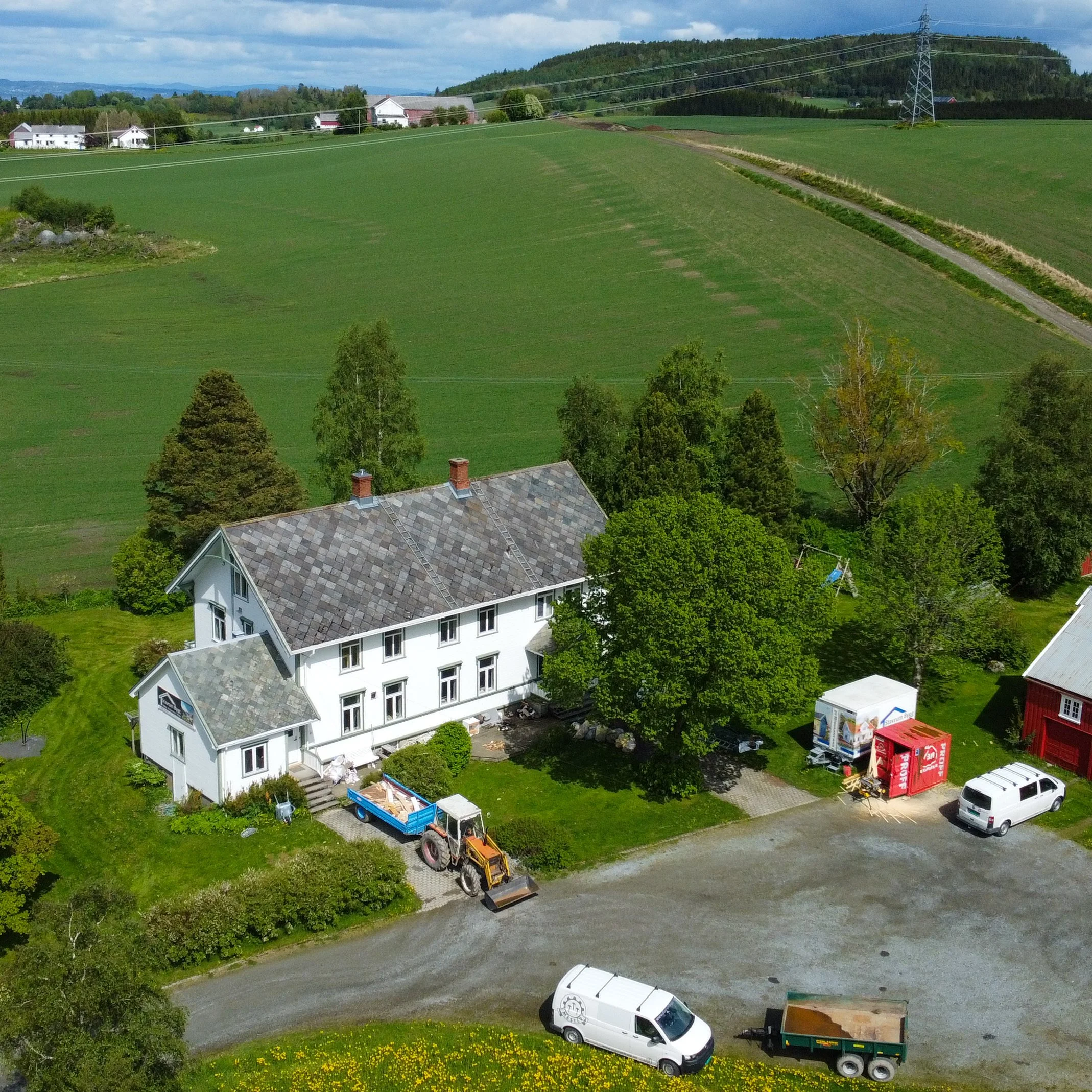Et hvitt hus med grått tak omgitt av grønne trær, biler og et traktor, med grønne åser i bakgrunnen.