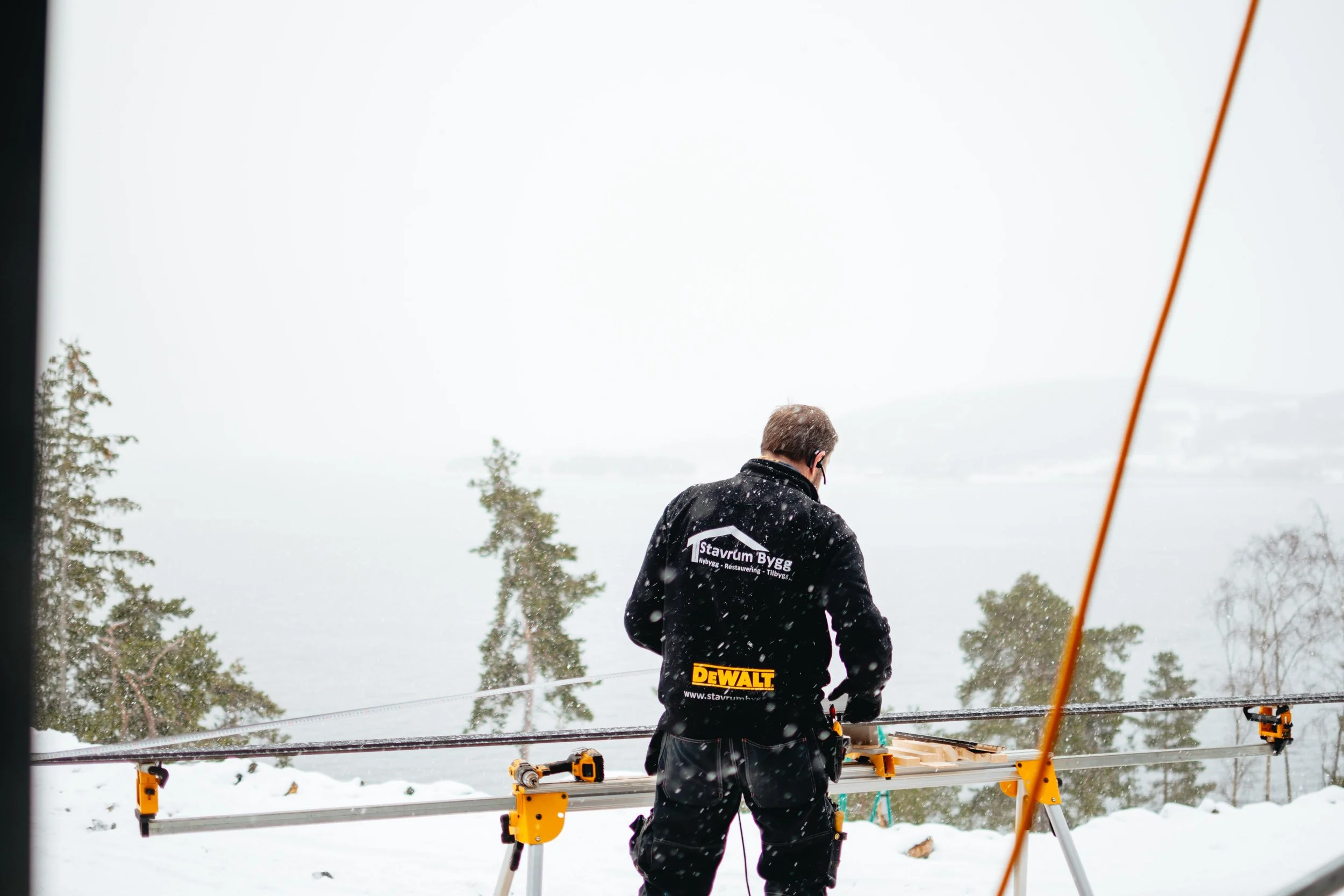 En person med svart jakke og arbeidsverktøy som står ute i snøen med et fjellandskap og trær i bakgrunnen, vintervær og snødekte områder