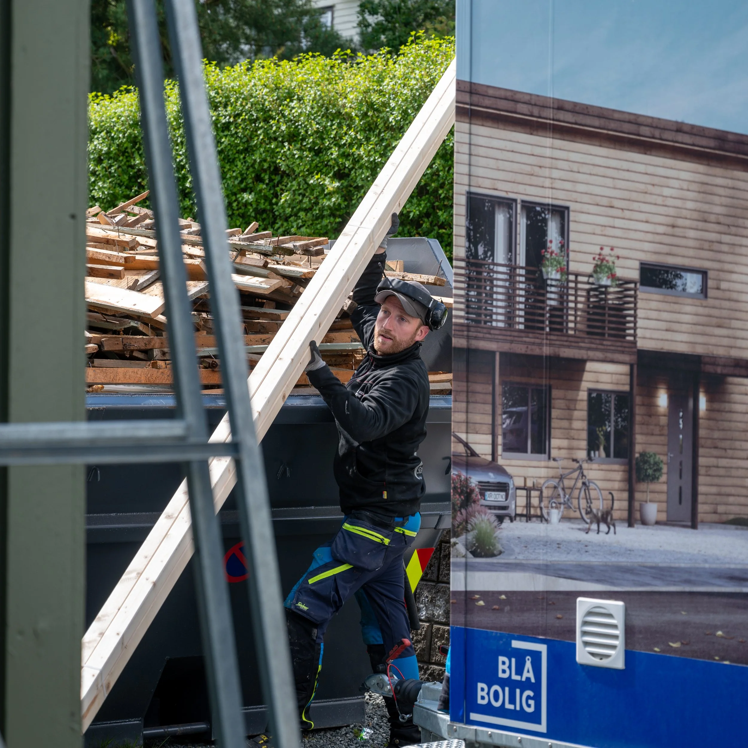 En mann i svart jakke og byxor med refleksprikker, med vernebriller og høretelefoner, jobber med treverk på en byggeplass. Det er en stor plakat av enebolig med trevegger og balkong på høyre side. På plakatens bunn står det "BLÅ BOLIG".