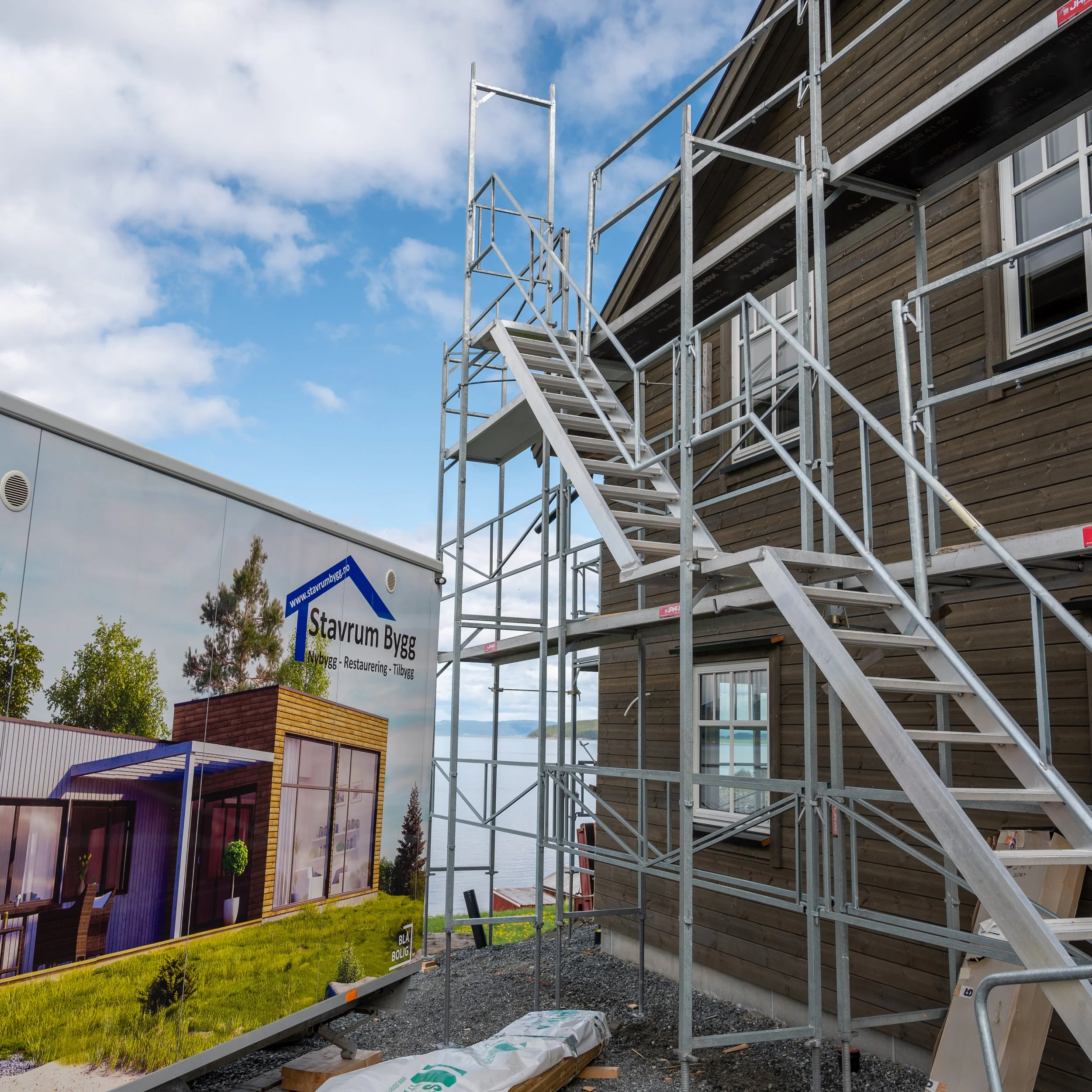 Byggearbeid med stillas foran en trekledning hus, plakat for Stavrum Bygg, som viser en modell av et ferdig hus, ute under en blå himmel med noen skyer.