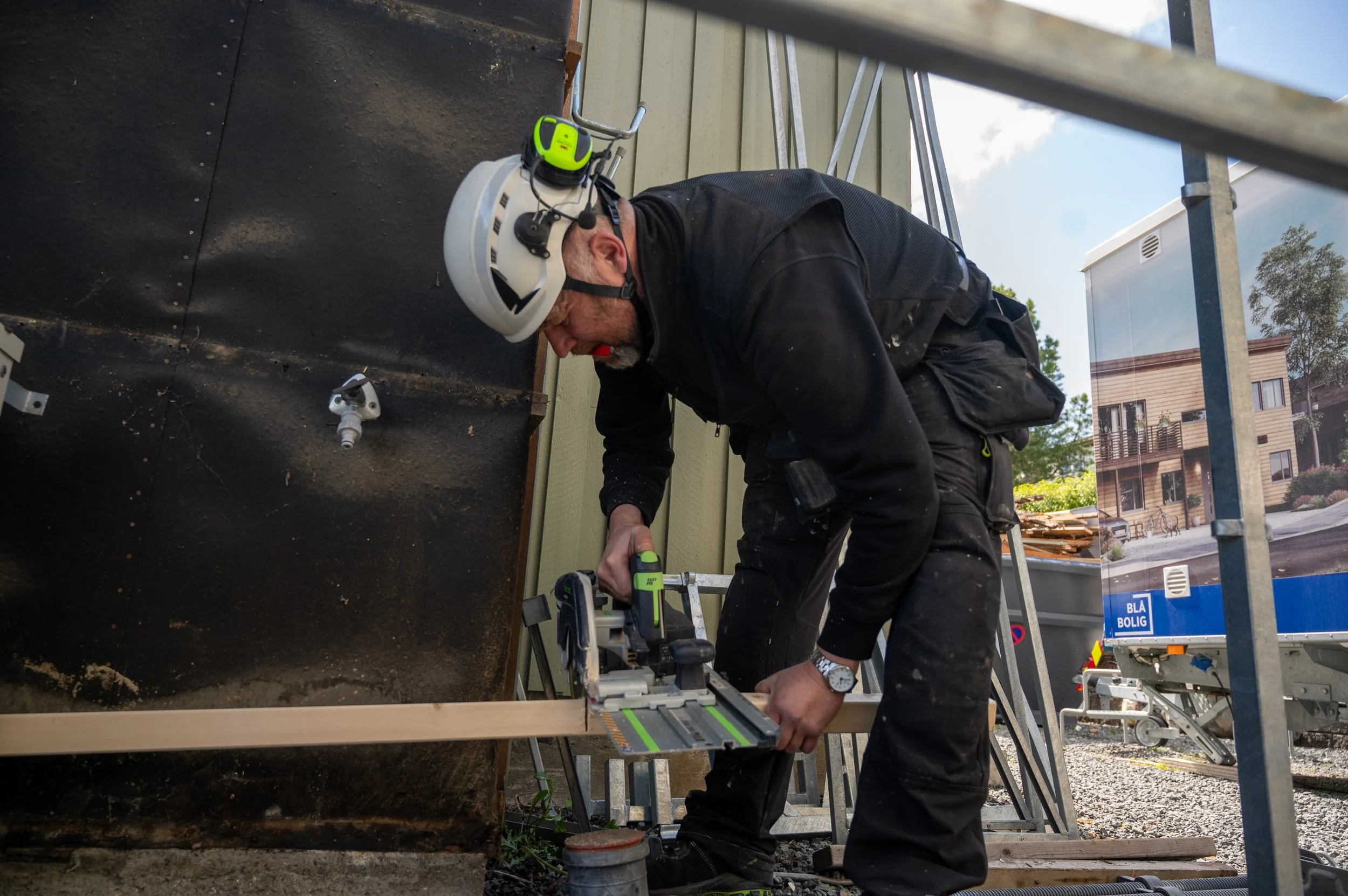 En mann med verneutstyr bruker en elektrisk sag på en byggeplass, jobber med en trelist.