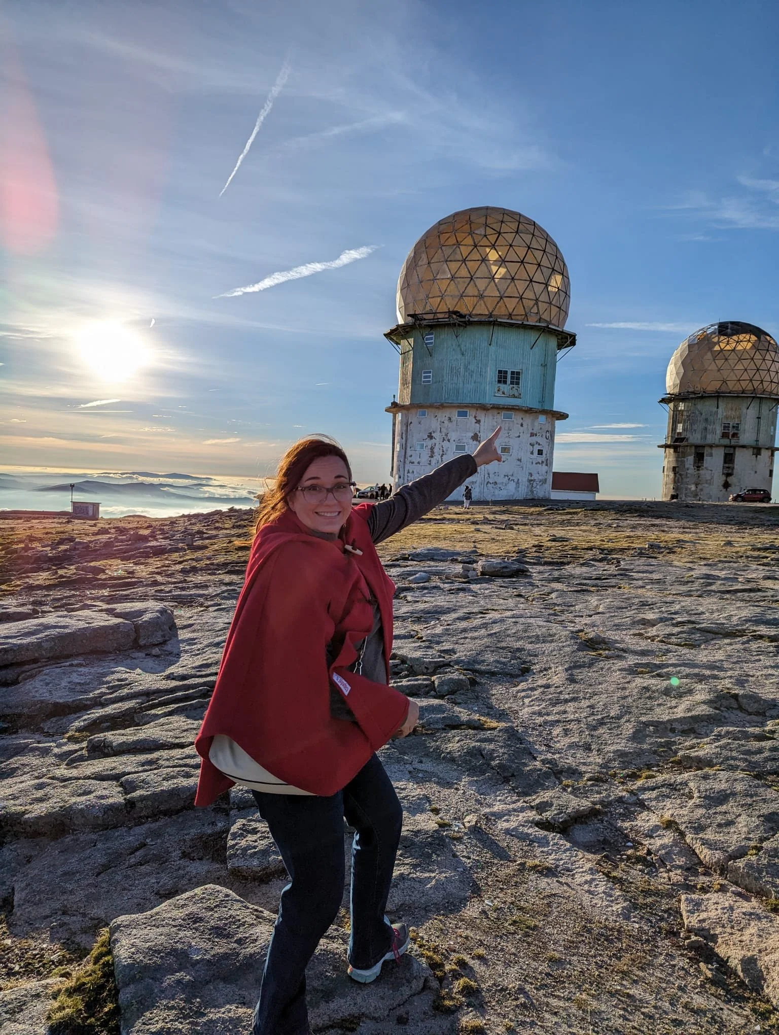 A woman in a red alentejo cape pointing at the highest point i mainland Portugal, the Torre (tower) in Serra da Estrela.