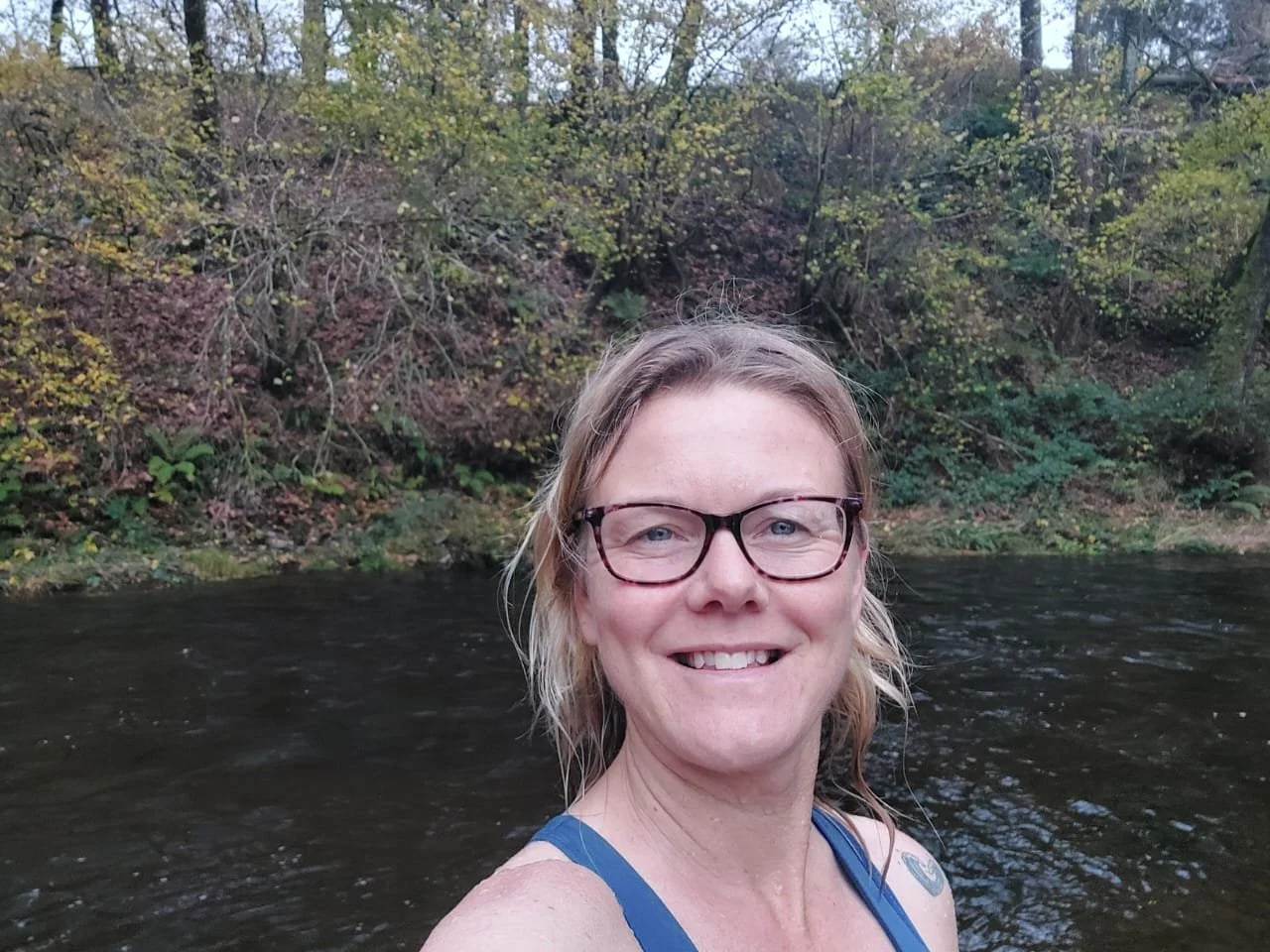 Sunrise over a partially frozen lake with rocks and small trees, surrounded by evergreen trees and snow-covered mountains.