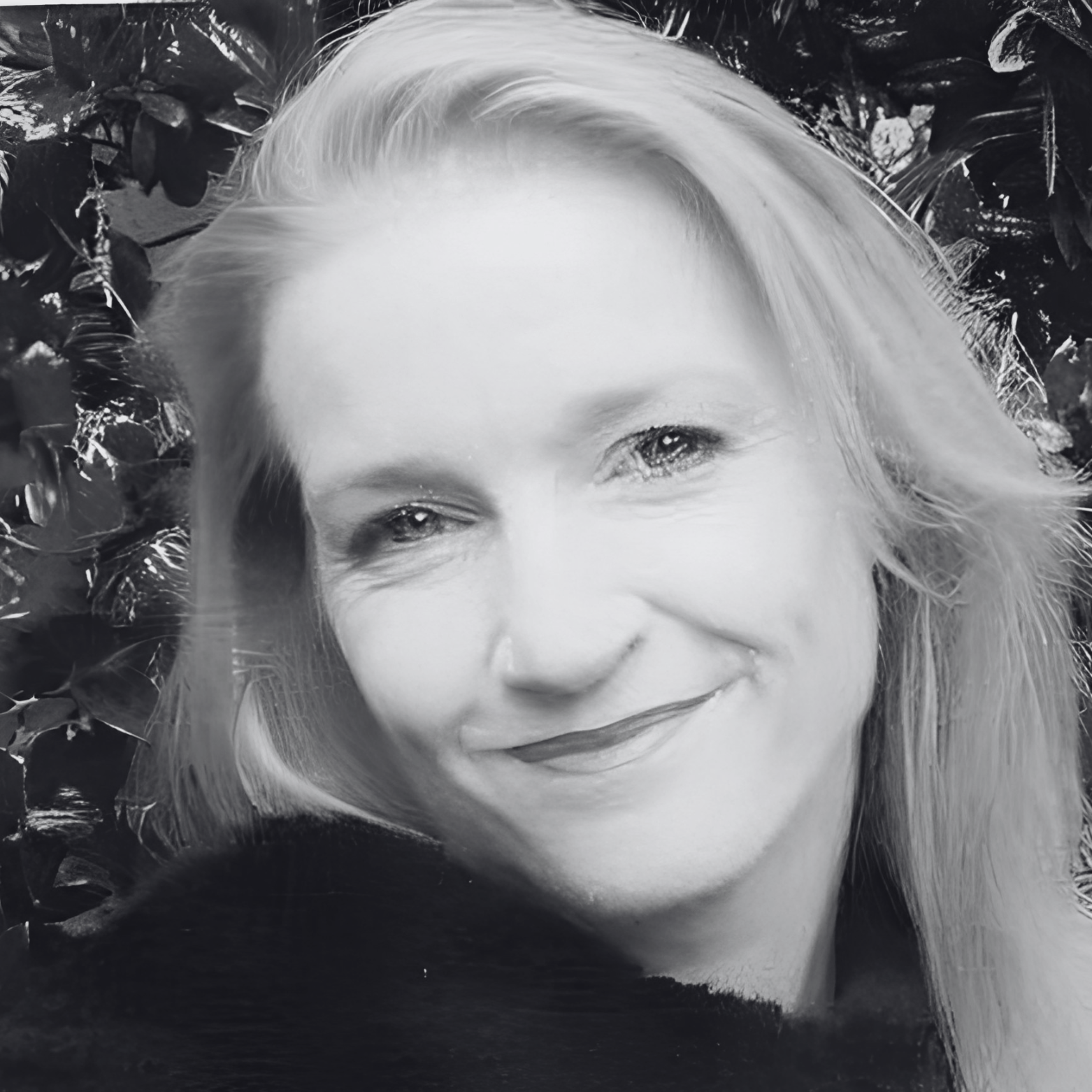 Black and white portrait of a woman with blonde hair, smiling, with leafy background.