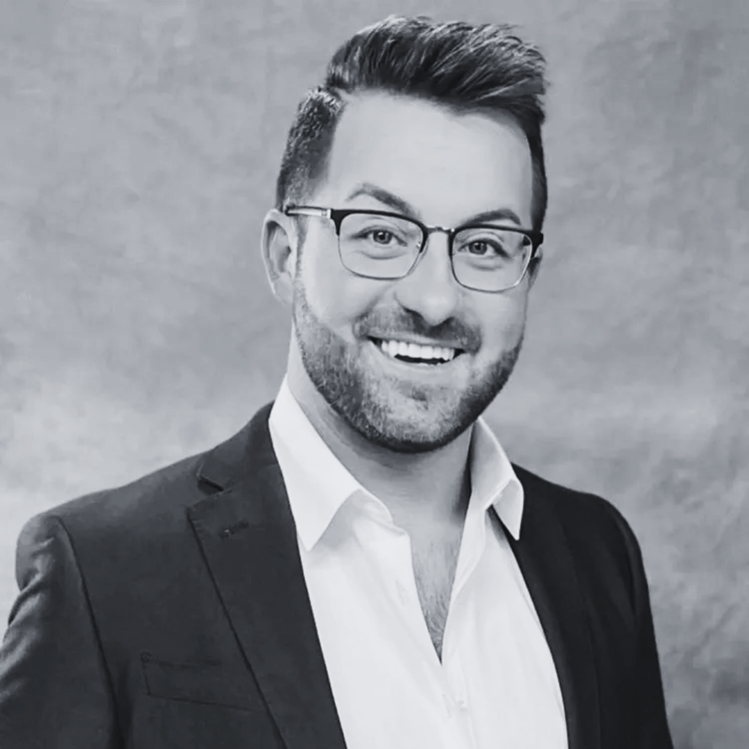 Black and white portrait of a smiling man with glasses, beard, and styled hair, wearing a suit jacket and white shirt.