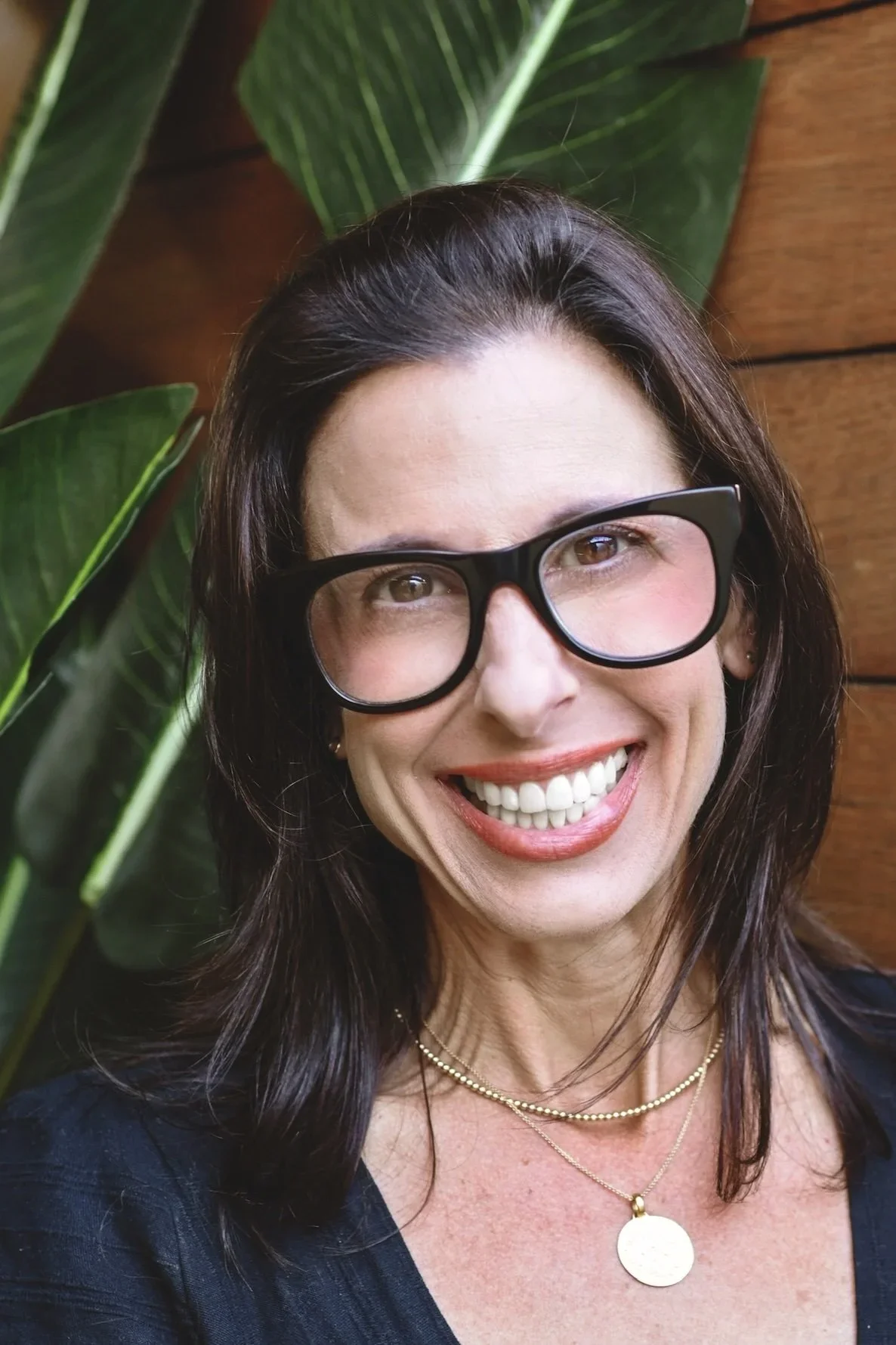 A smiling woman with dark hair, wearing large black glasses and a gold necklace, standing in front of green plants and a wooden wall.