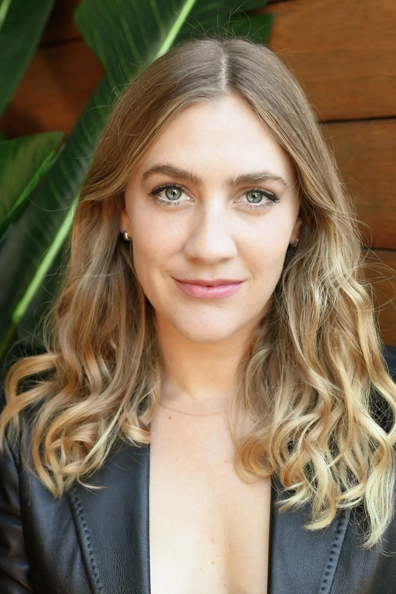 A young woman with blonde, wavy hair, wearing a black leather jacket and pearl earrings, looking at the camera with a slight smile. Green plants and a wooden wall are in the background.