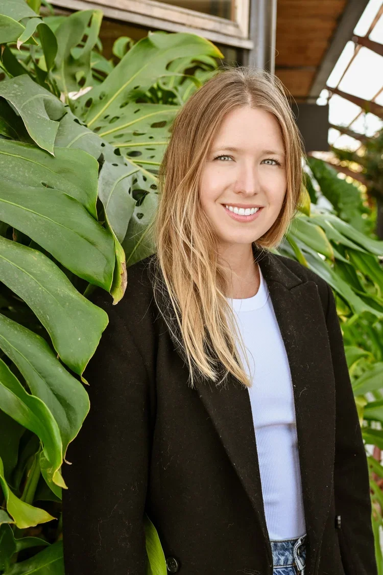 Young woman with blonde hair smiling, standing near green large leafy plants in a greenhouse or garden.