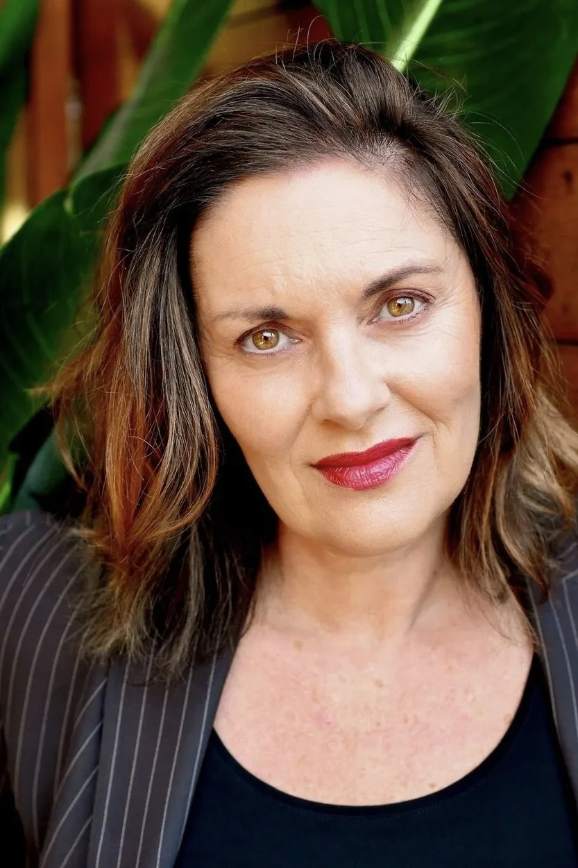 A woman with shoulder-length wavy brown hair, wearing a dark pinstripe blazer and red lipstick, standing outdoors in front of large green leaves.
