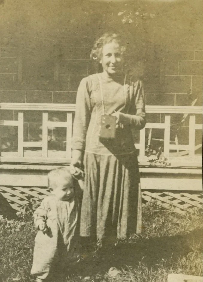 Black and white photo of a woman holding an old Brownie camera in one hand against her chest. A  toddler is standing beside her holding her hand. They are in front of the John Brown House.