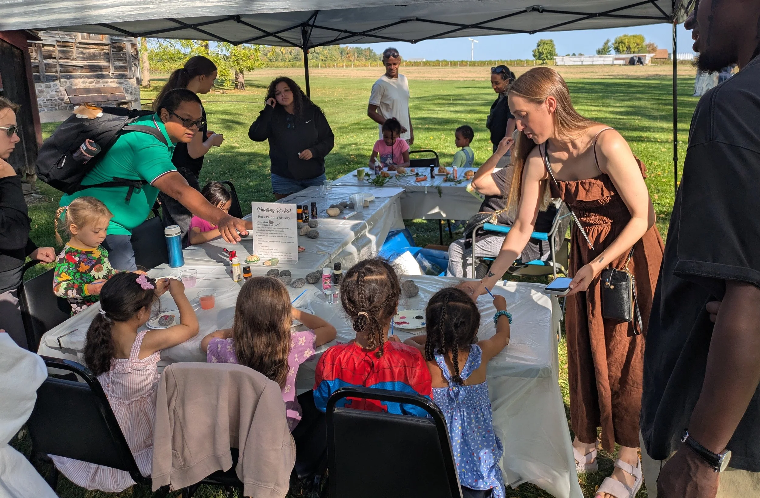 A large group of of children and their families gather around tables of crafts. They are outside under a tent.