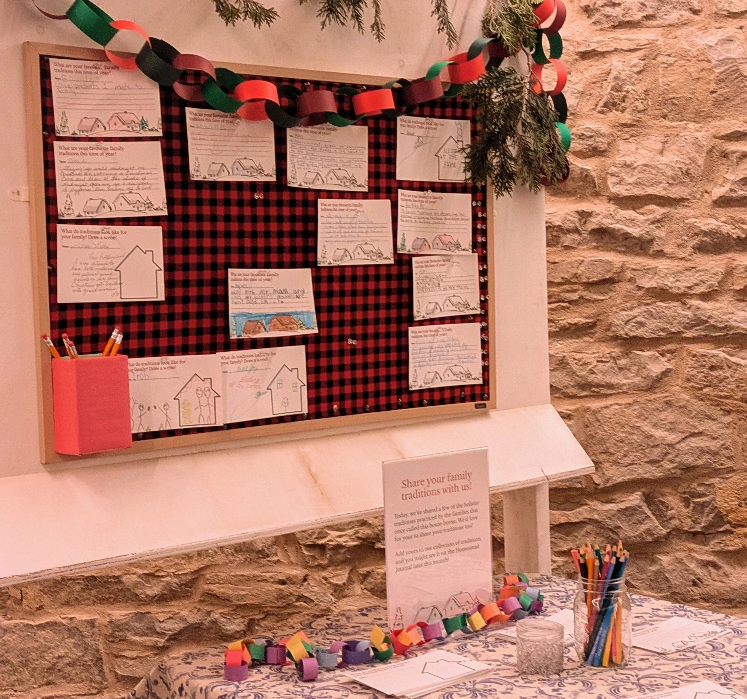 A wooden display panel is wrapped in red and black checkered fabric and draped with paper chains. The panel is covered in hand-written and hand-drawn notes highlighting submitters' family traditions around the holidays.