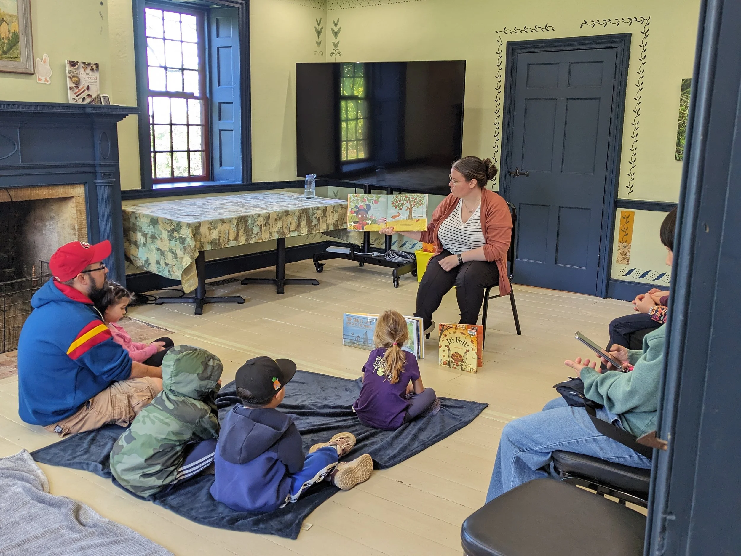 Children sit on blankets, listening to a woman as she reads a storybook in her hand.