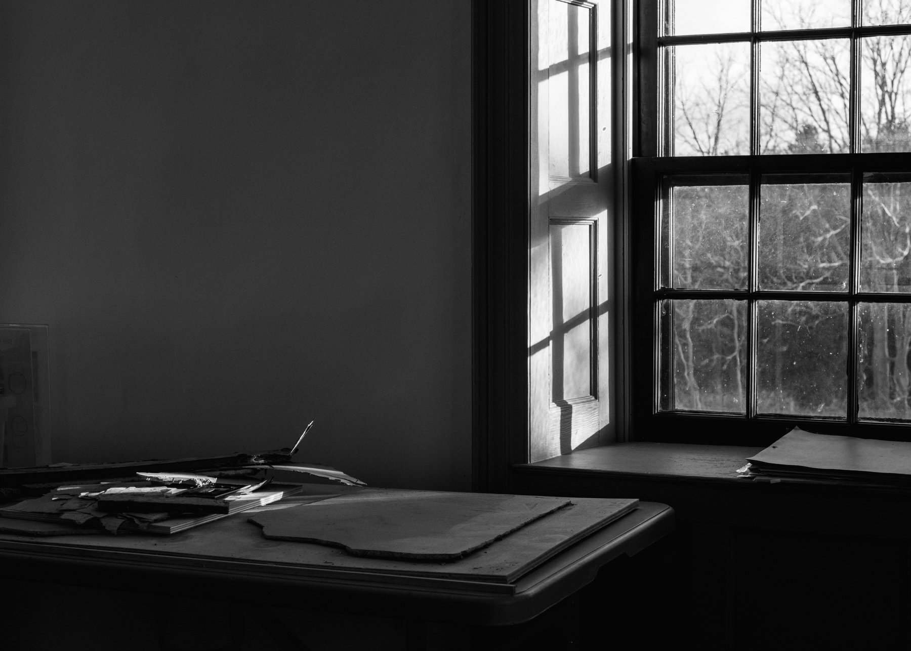 Moody black and white photograph, with sunshine filtering through an old window casting shadows over a cluttered tabletop.