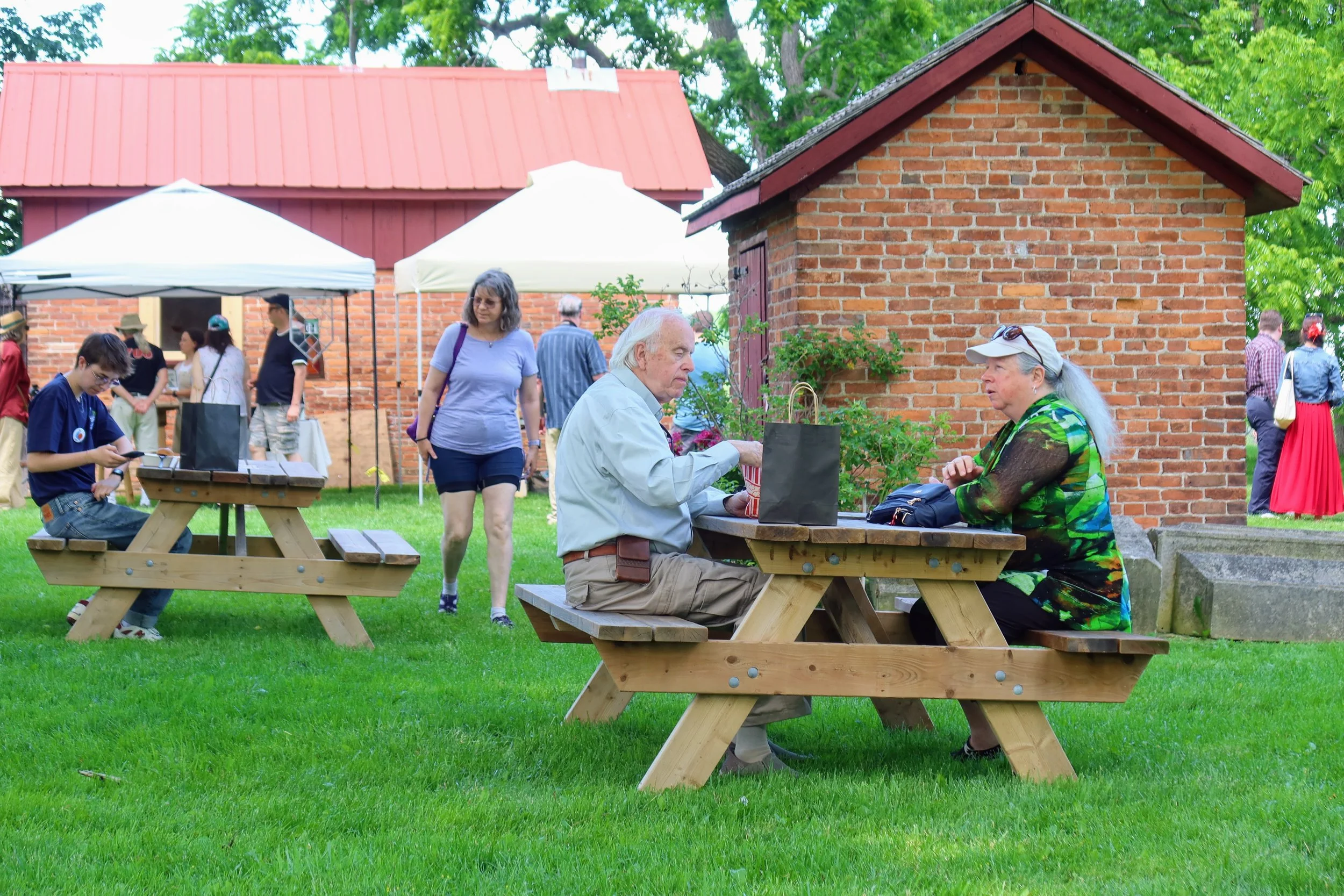 Visitors eat at picnic tables outside. Behind them is the red brick of a historic smokehouse and dairy as well as tents with heritage demonstrators working.