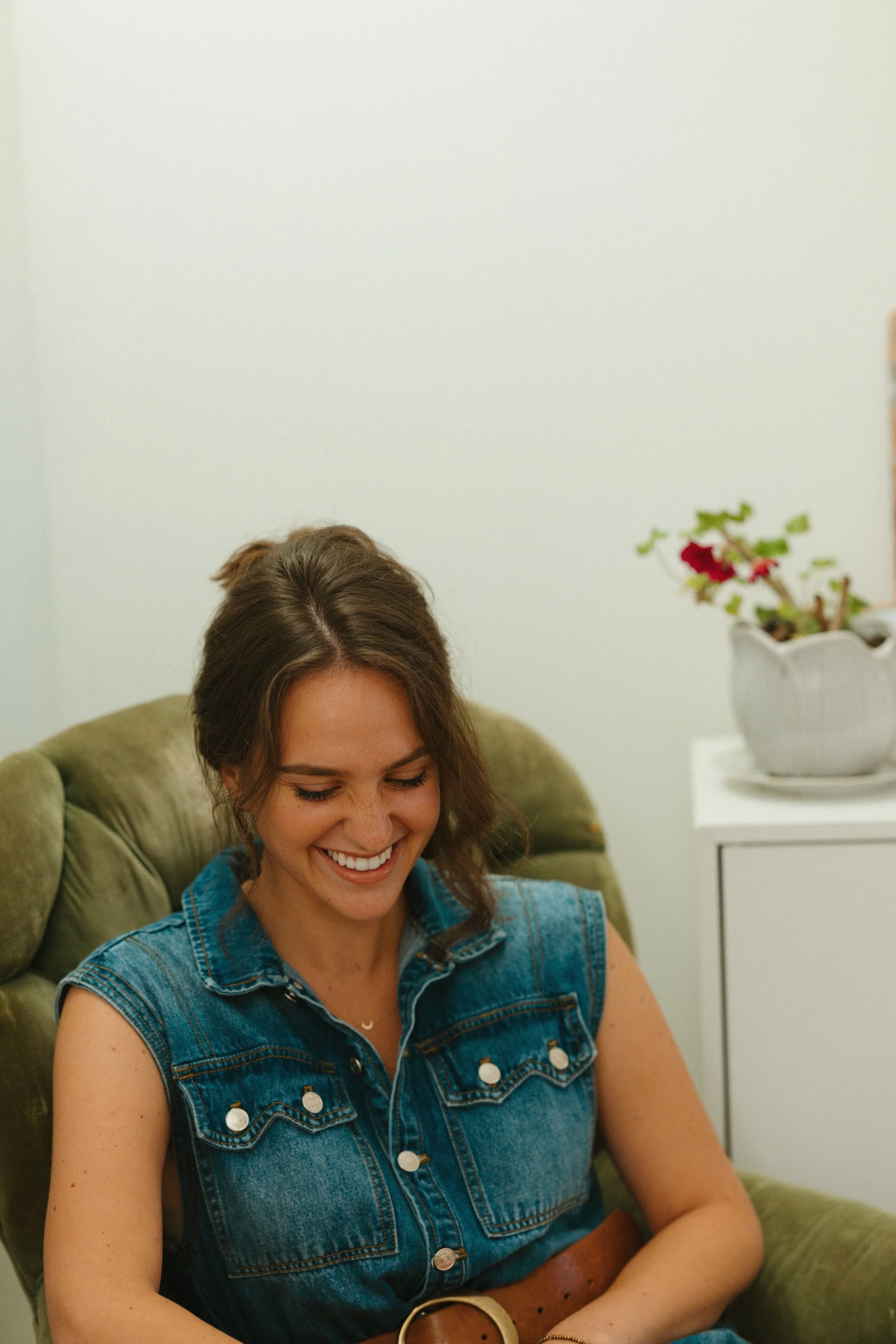 A young woman smiles as she sits in a olive green cushioned armchair.