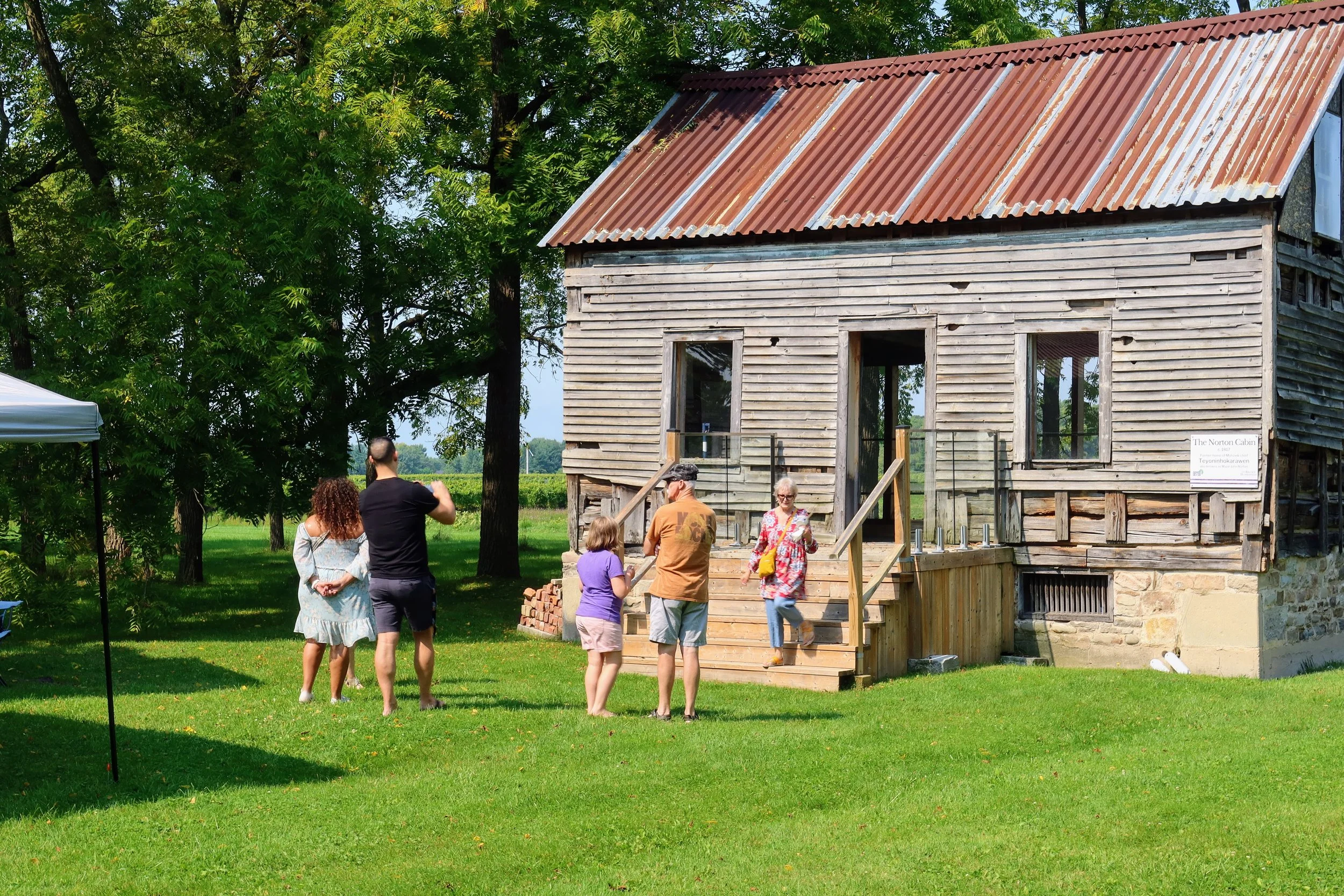 Visitors chat around a wooden cabin with a metal roof. The cabin is surrounded by trees.