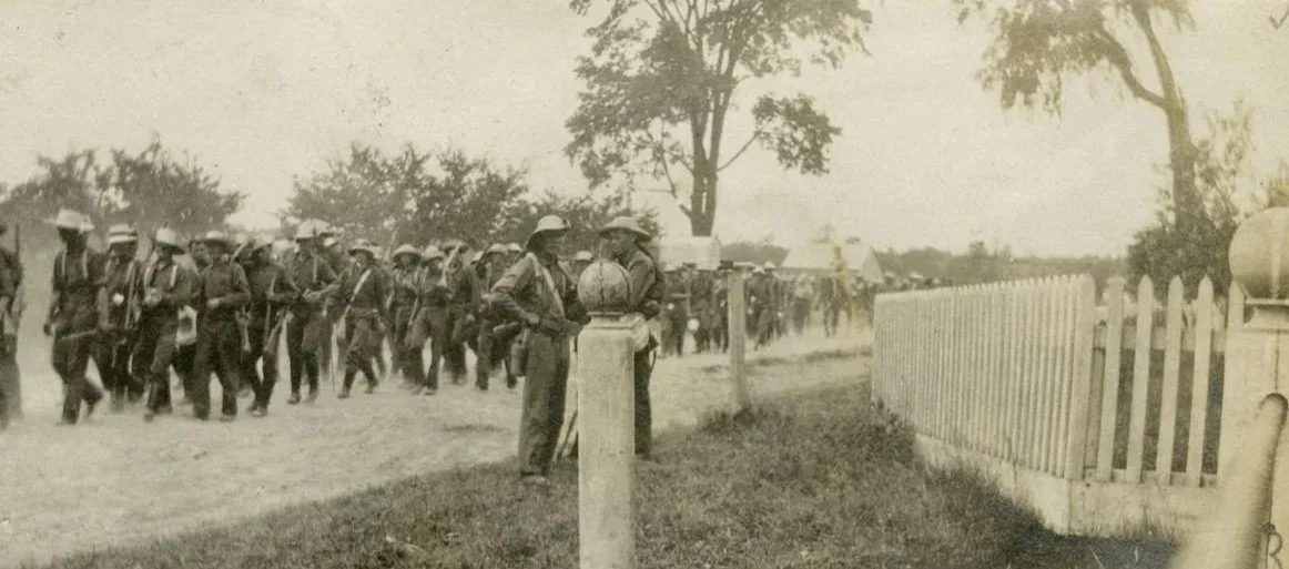 Soldiers march along a dirt road, carrying long guns and wearing wide rimmed hats. The photo is taken behind a white fence.