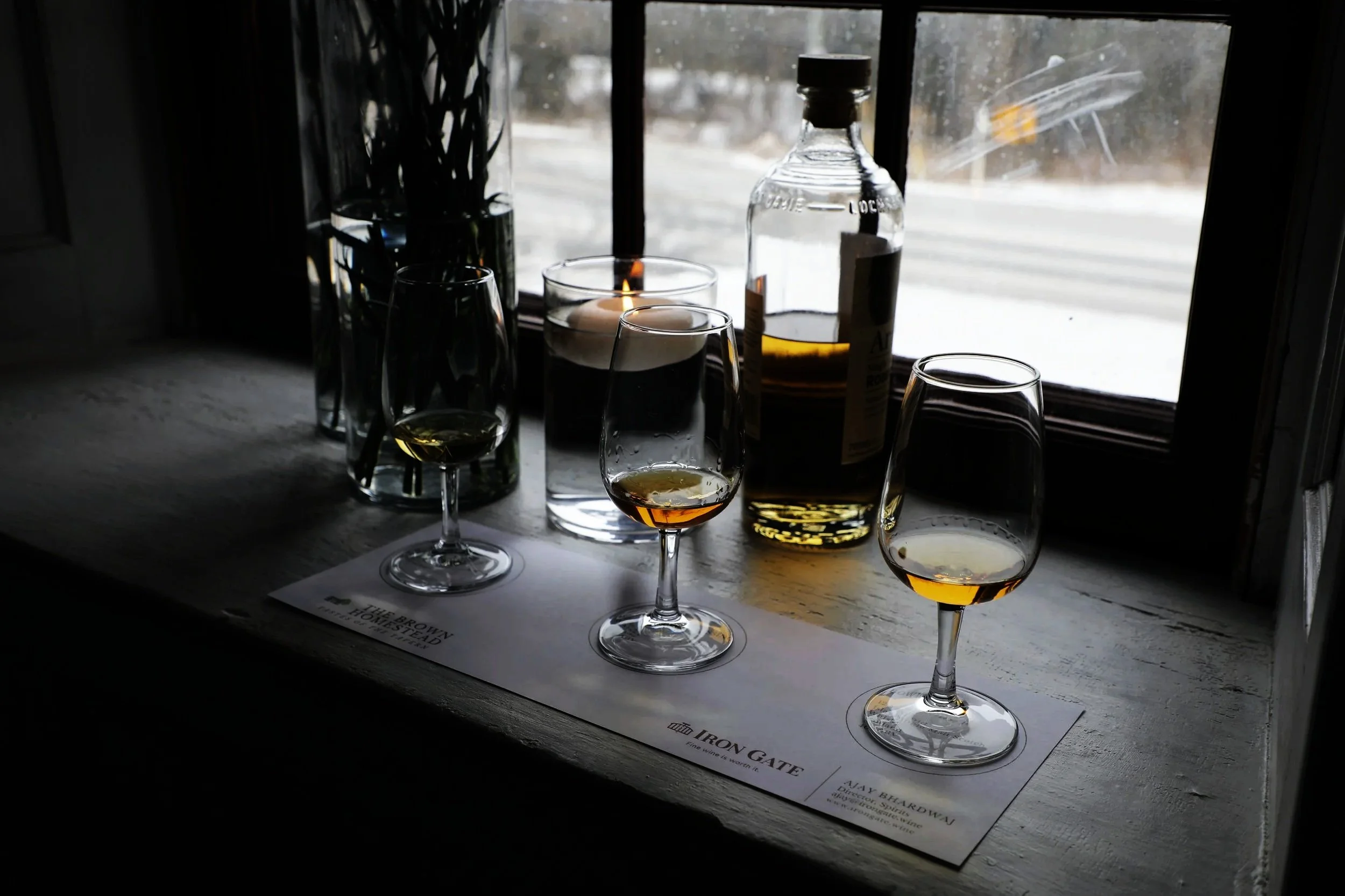 Three drams of whisky sit on a window sill, with a candle floating in a vase of water and a half open bottle of whisky in the background.