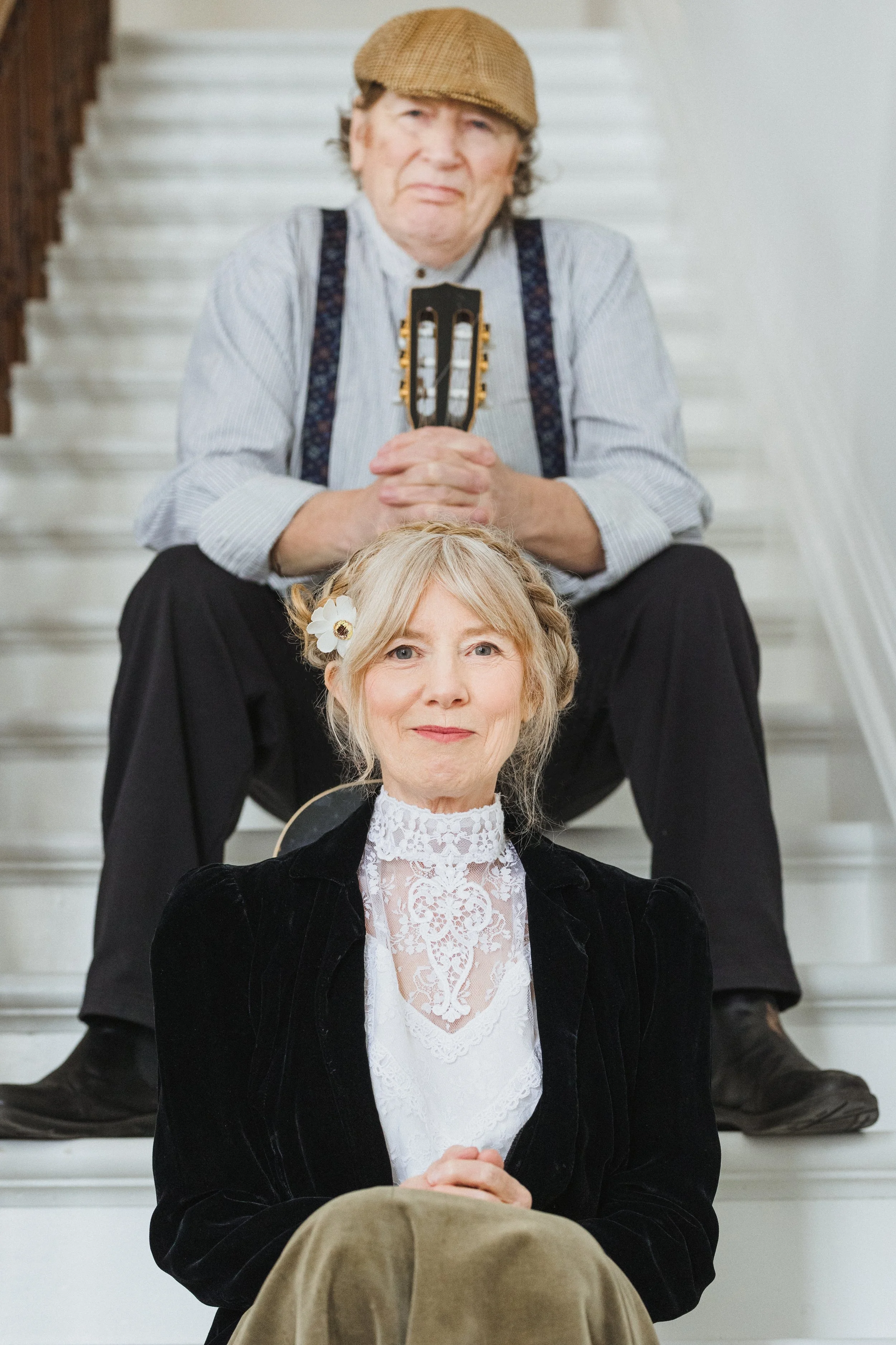 An older women wears a mischievous smile as she sits on the steps of a tall staircase, a man sits on a step behind her holding a guitar. They are dressed in Edwardian costume of the 1910s.