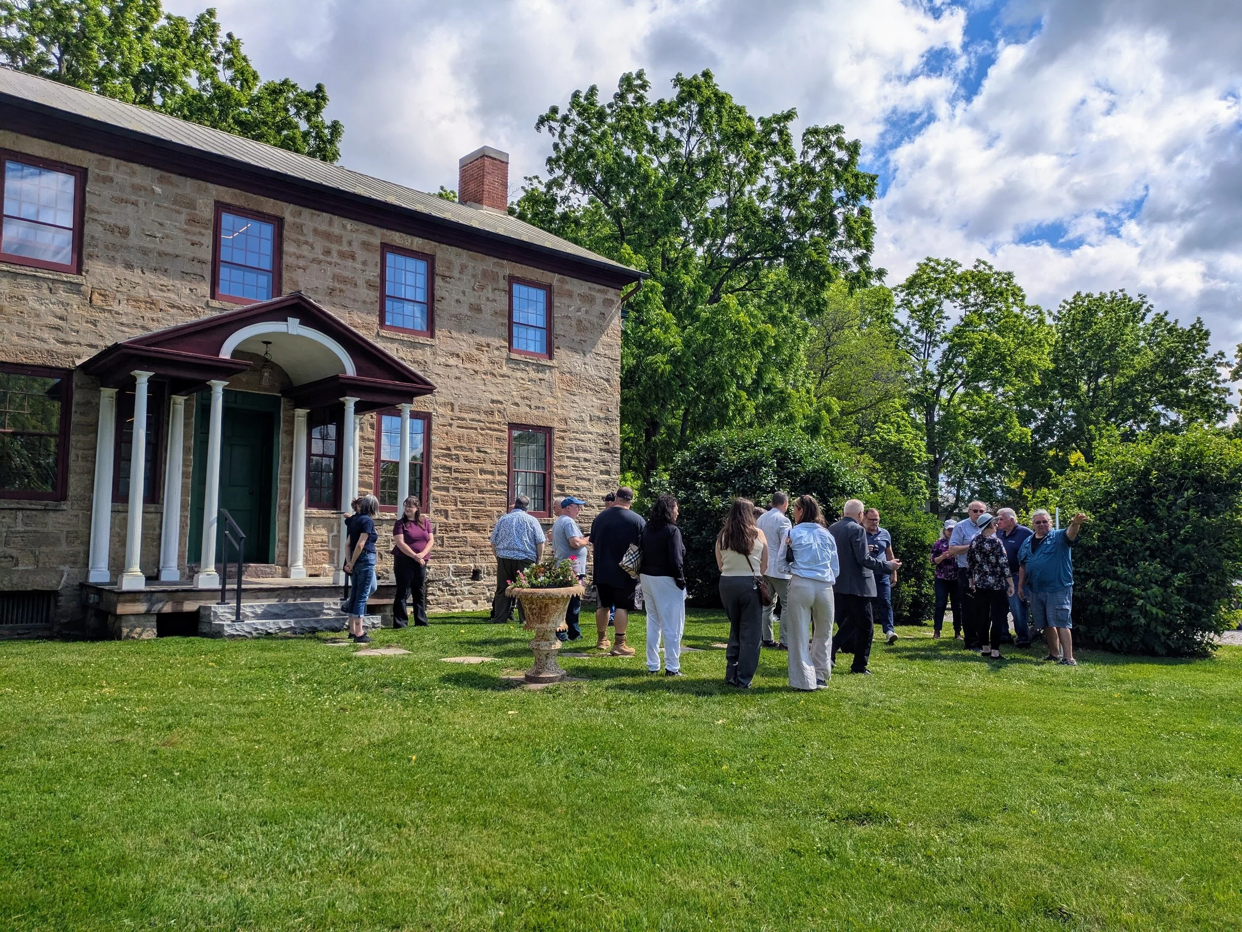 A group gathers in front of a two storey stone house, built in the Georgian style.