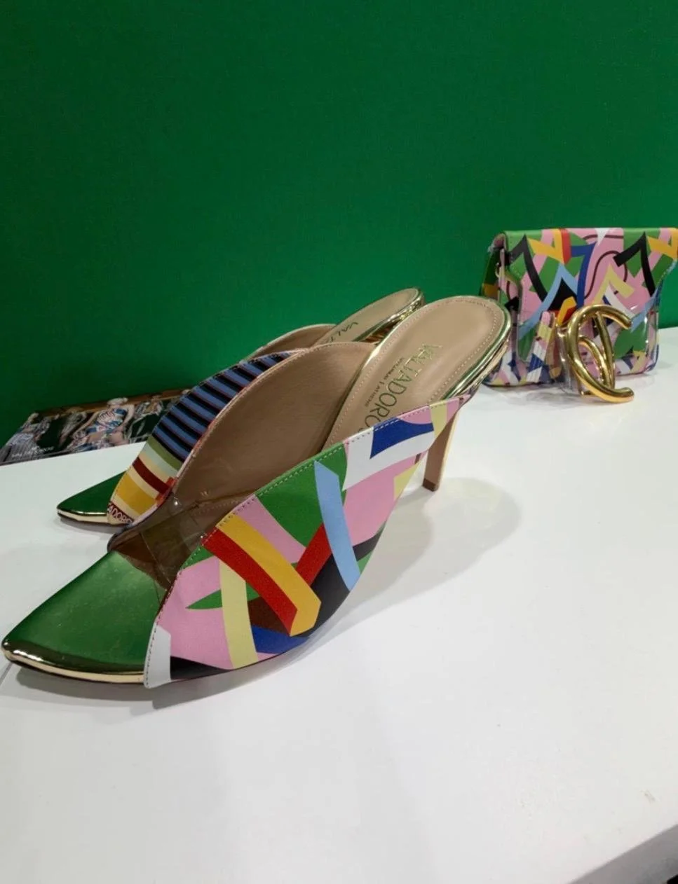 Une paire de talons hauts colorés, une pochette avec un design abstrait et un logo doré posé sur une table contre un fond vert.