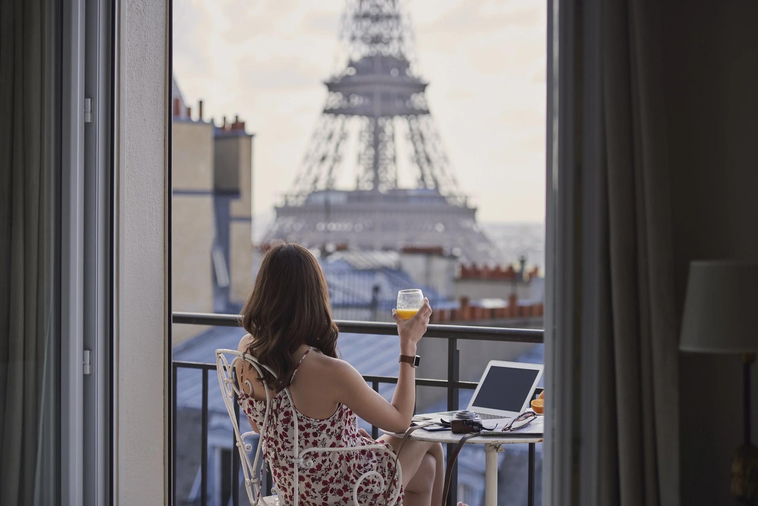 Une femme assise sur un balcon, tenant un verre orange, avec une tablette sur la table devant elle, face à la Tour Eiffel à Paris, vue depuis une fenêtre
