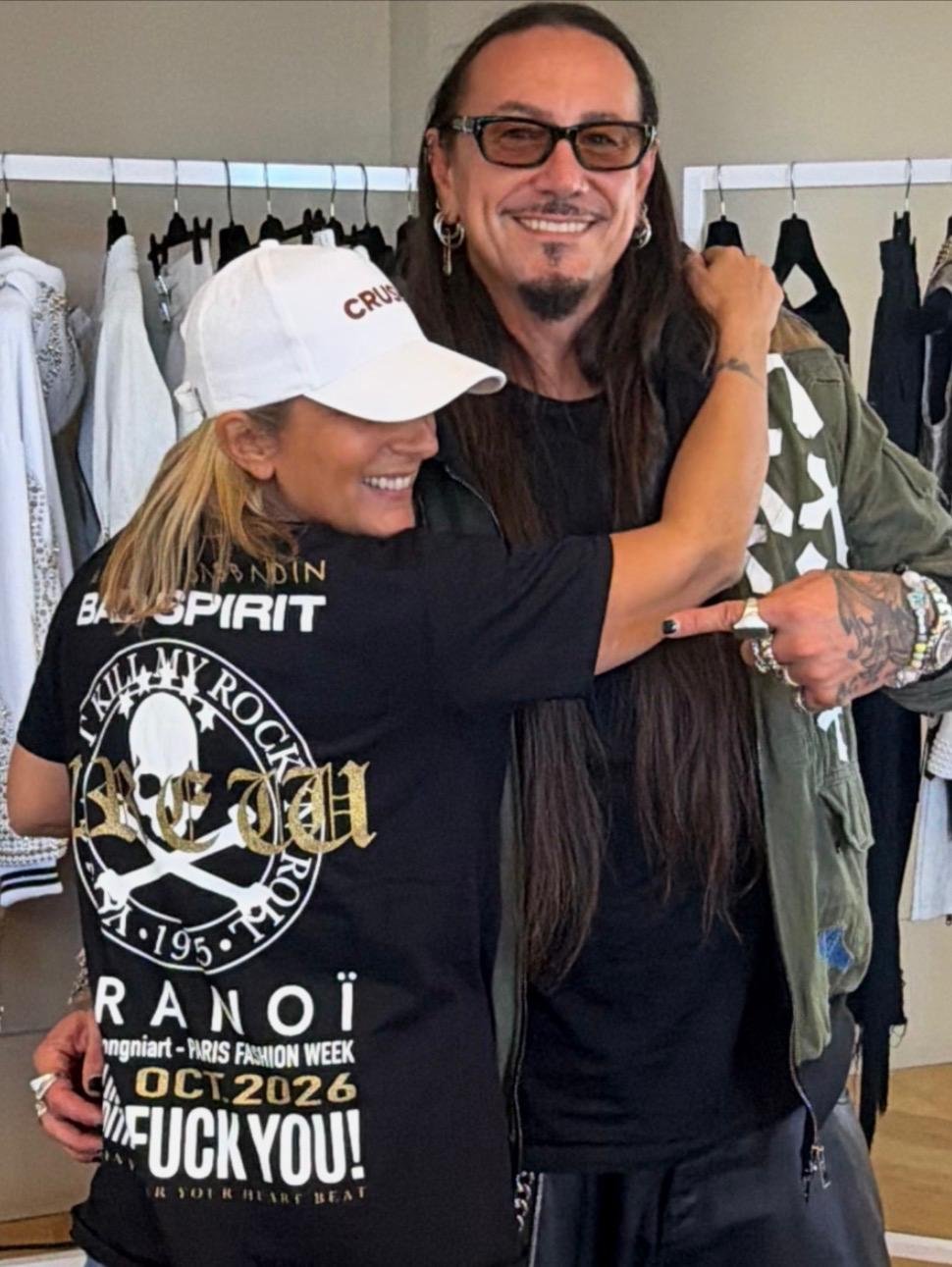 Deux personnes souriantes, une femme portant un t-shirt noir avec des inscriptions et un homme portant des vêtements à motifs, posant ensemble en magasin de vêtements.