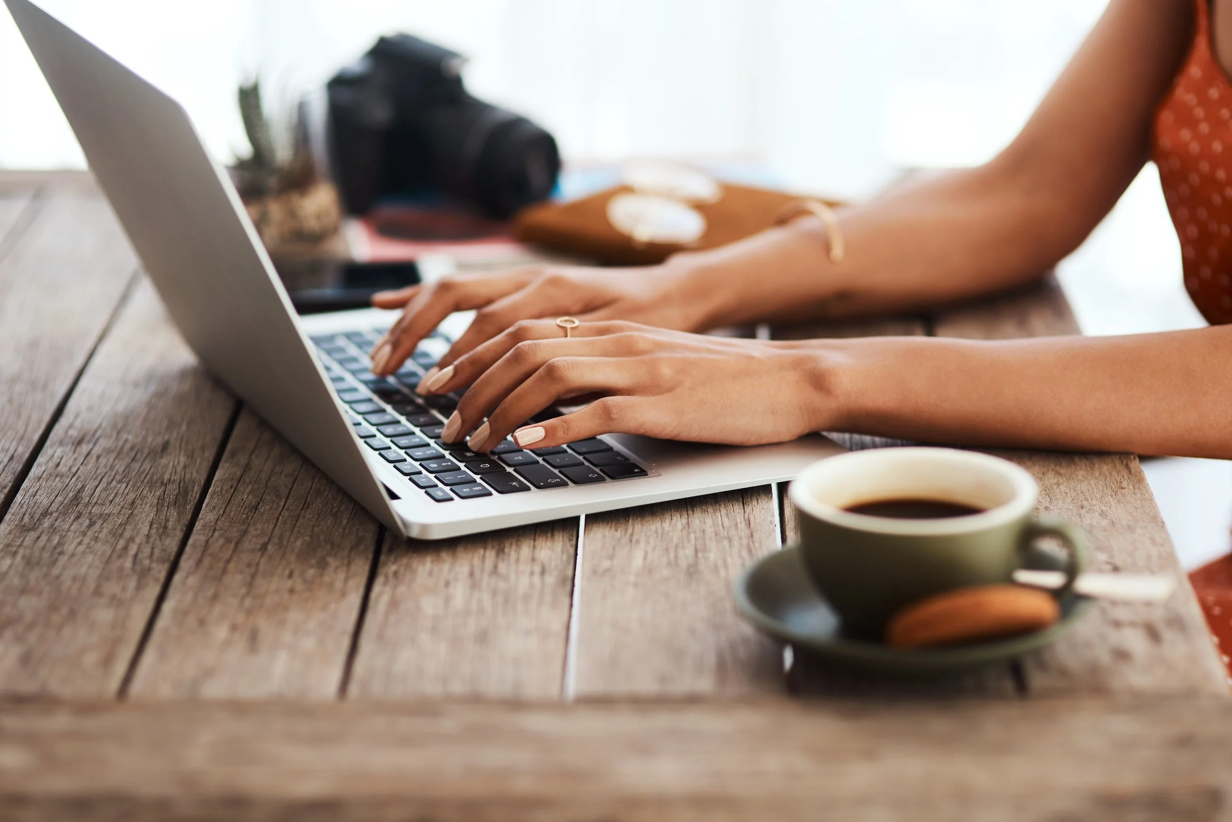 Personne tapant sur un clavier d'ordinateur portable sur une table en bois, à côté d'une tasse de café et d'un appareil photo en arrière-plan.