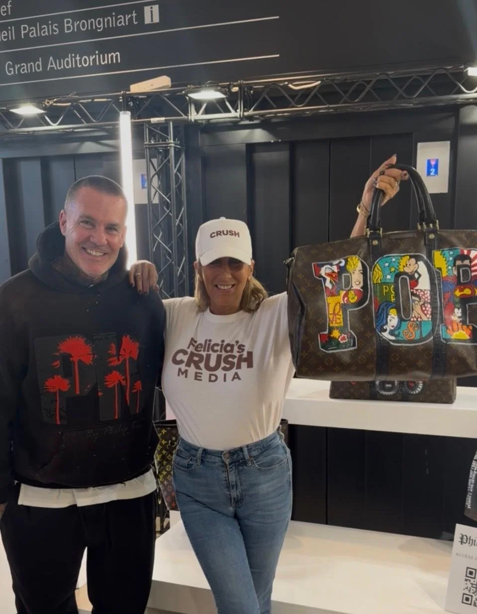 Deux personnes souriantes posent pour une photo, une femme tient un grand sac aux lettres colorées. Elles sont dans un lieu avec des panneaux d'information en français et en anglais, notamment indiquant le Palais Brongiart et le Grand Auditorium.
