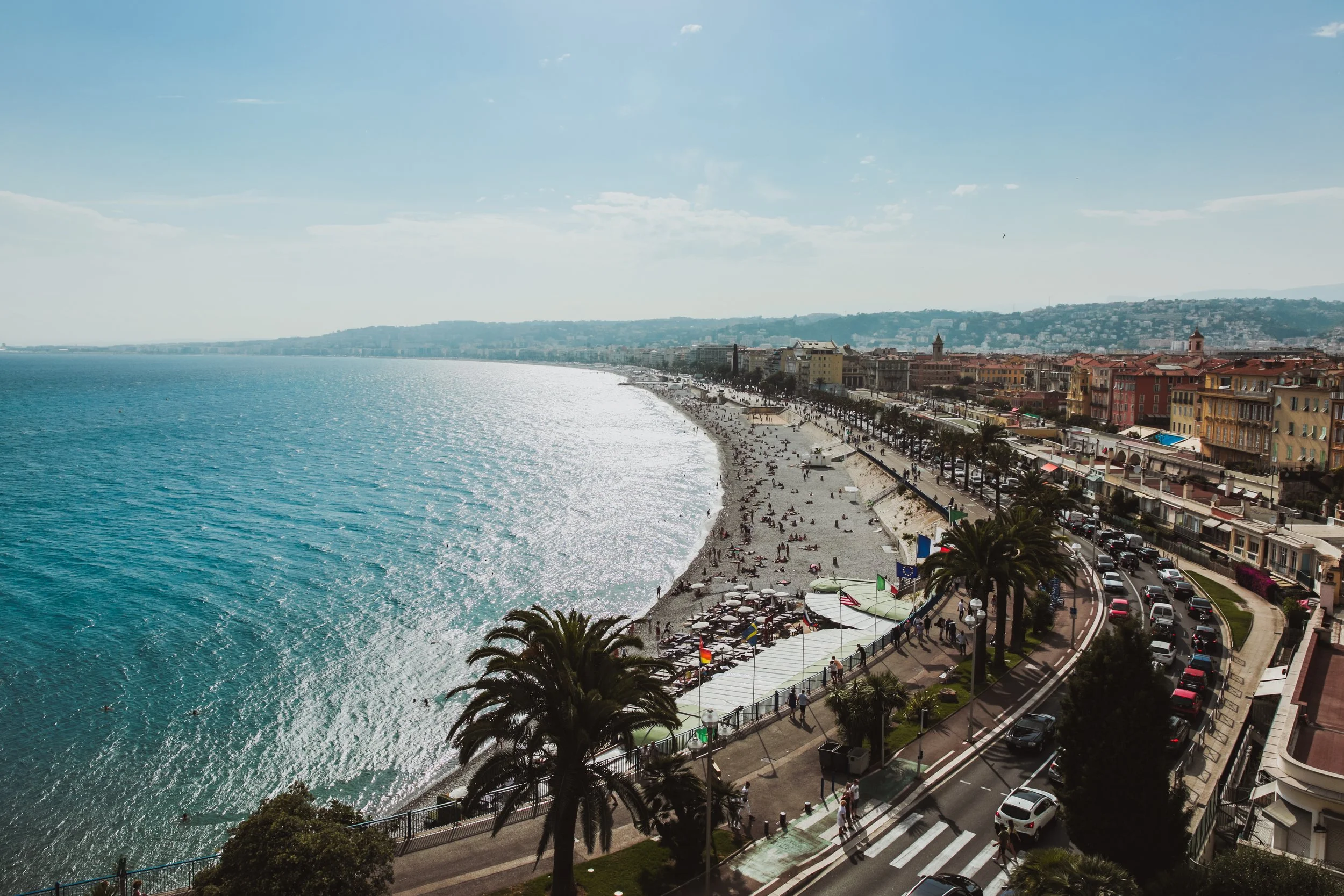 Vue panoramique d'une plage animée avec des parasols et des gens qui se détendent, bordant une promenade avec des bâtiments colorés et des palmiers, dans une ville côtière en journée ensoleillée.